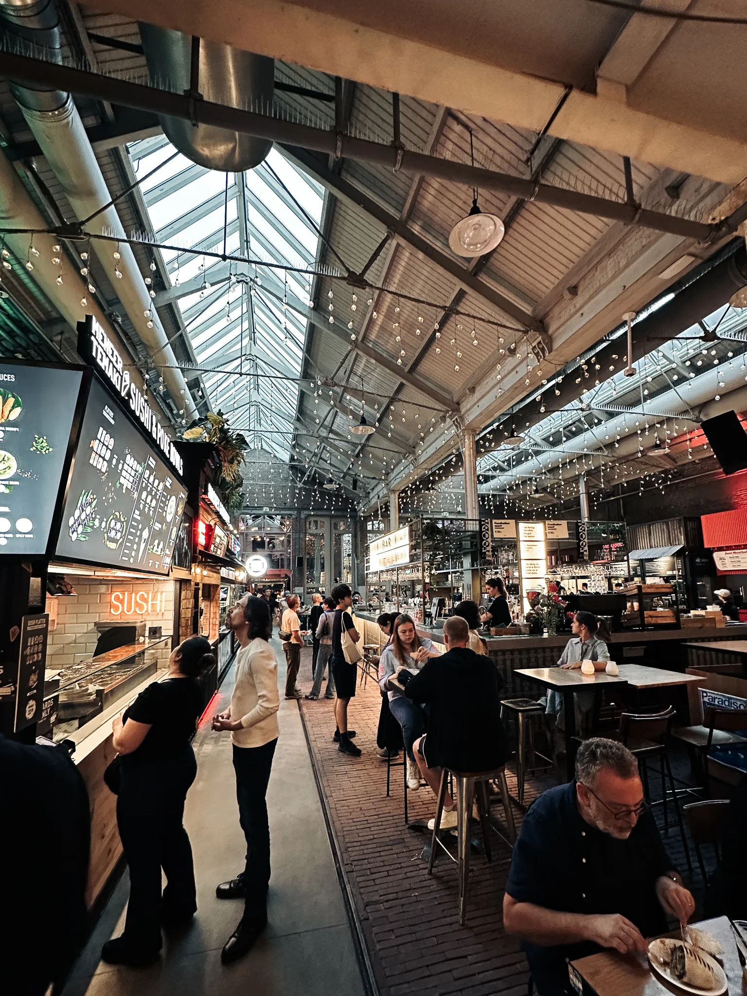 Inside the Foodhallen - steel trusses and a glazed pitched roof, strings of bulbs over the food stalls, a sushi counter lit at one end and people eating at high tables down the aisle