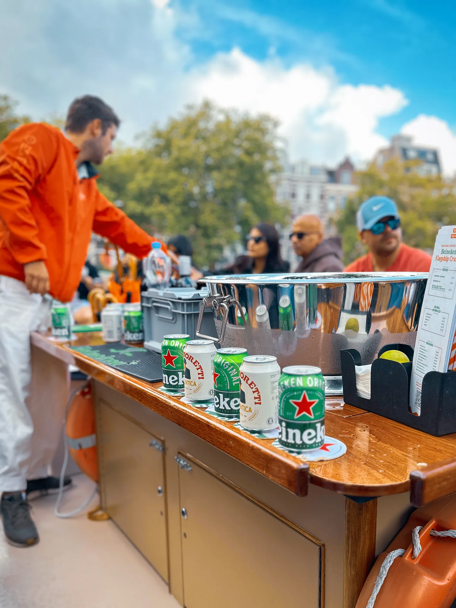 The bar of a canal cruise boat - cold Heineken and Moretti cans lined along the varnished counter beside a steel ice bucket, a crew member in an orange jacket reaching past and passengers in sunglasses behind