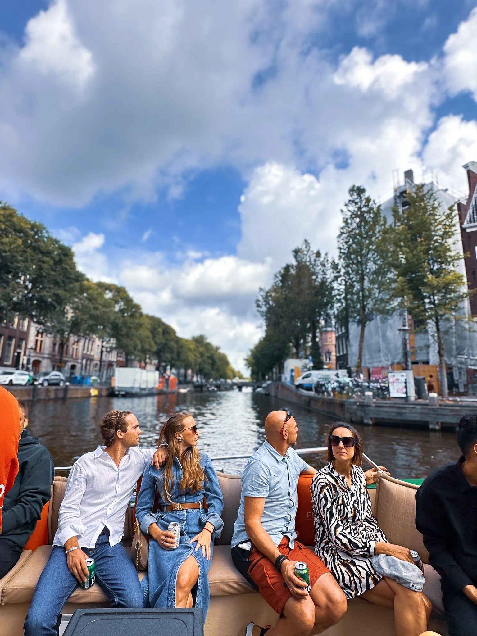 Four passengers on the padded bench of an open canal boat, drinks in hand and all looking off the same side, the canal walls and plane trees running away behind them under towering white cloud