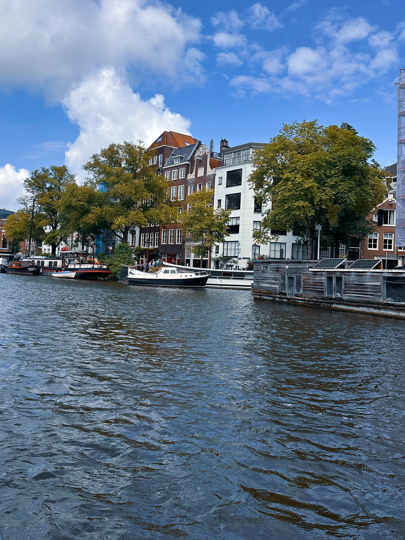 The boat swings out into the Amstel - chop on the surface, houseboats and a white motor cruiser moored along the far bank below brick gables and a white modern block, trees between them