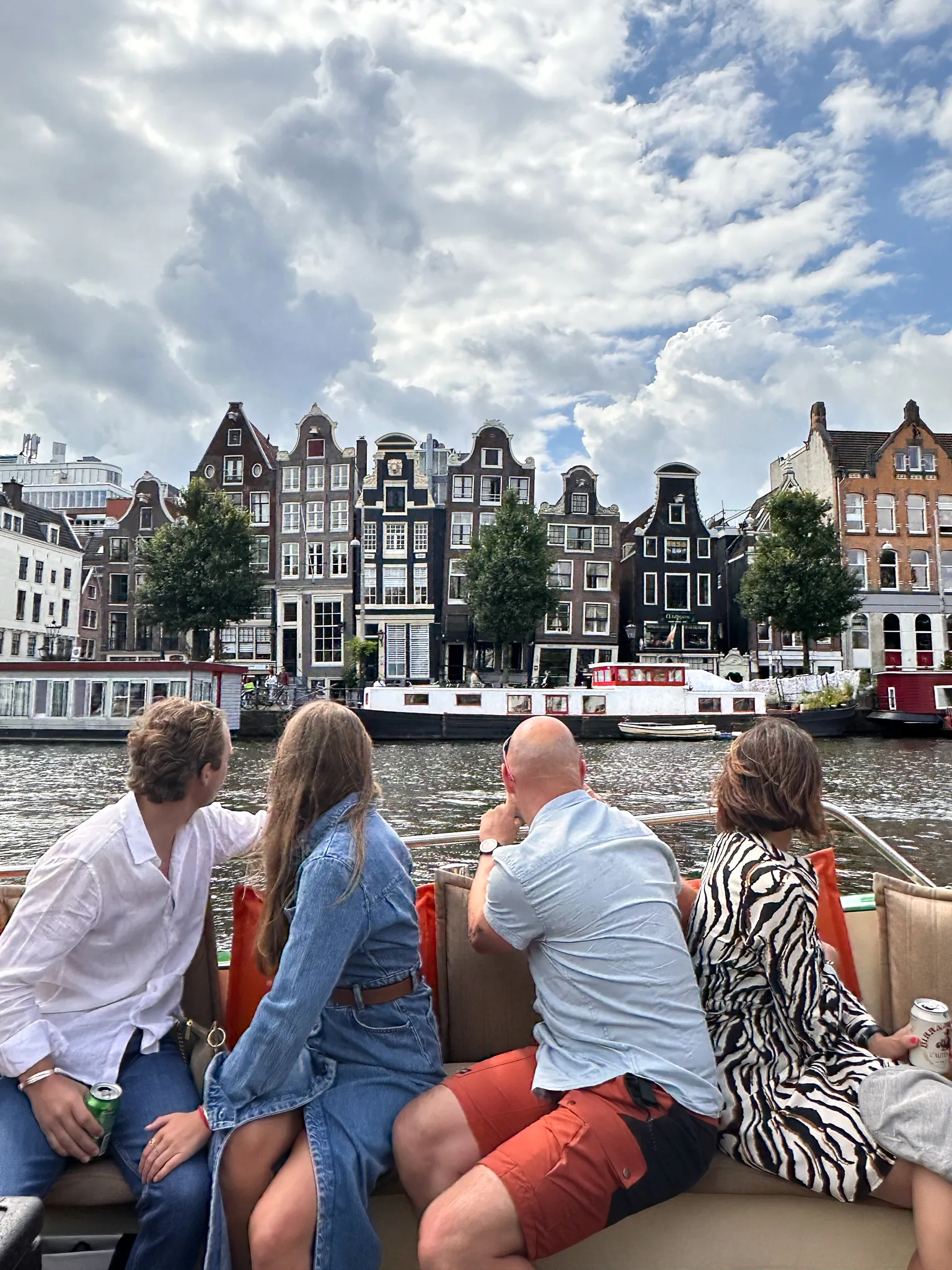 Seen from behind the bench, four passengers watch the leaning gabled houses on the Amstel slide past, each at its own angle, with houseboats moored along the quay in front of them