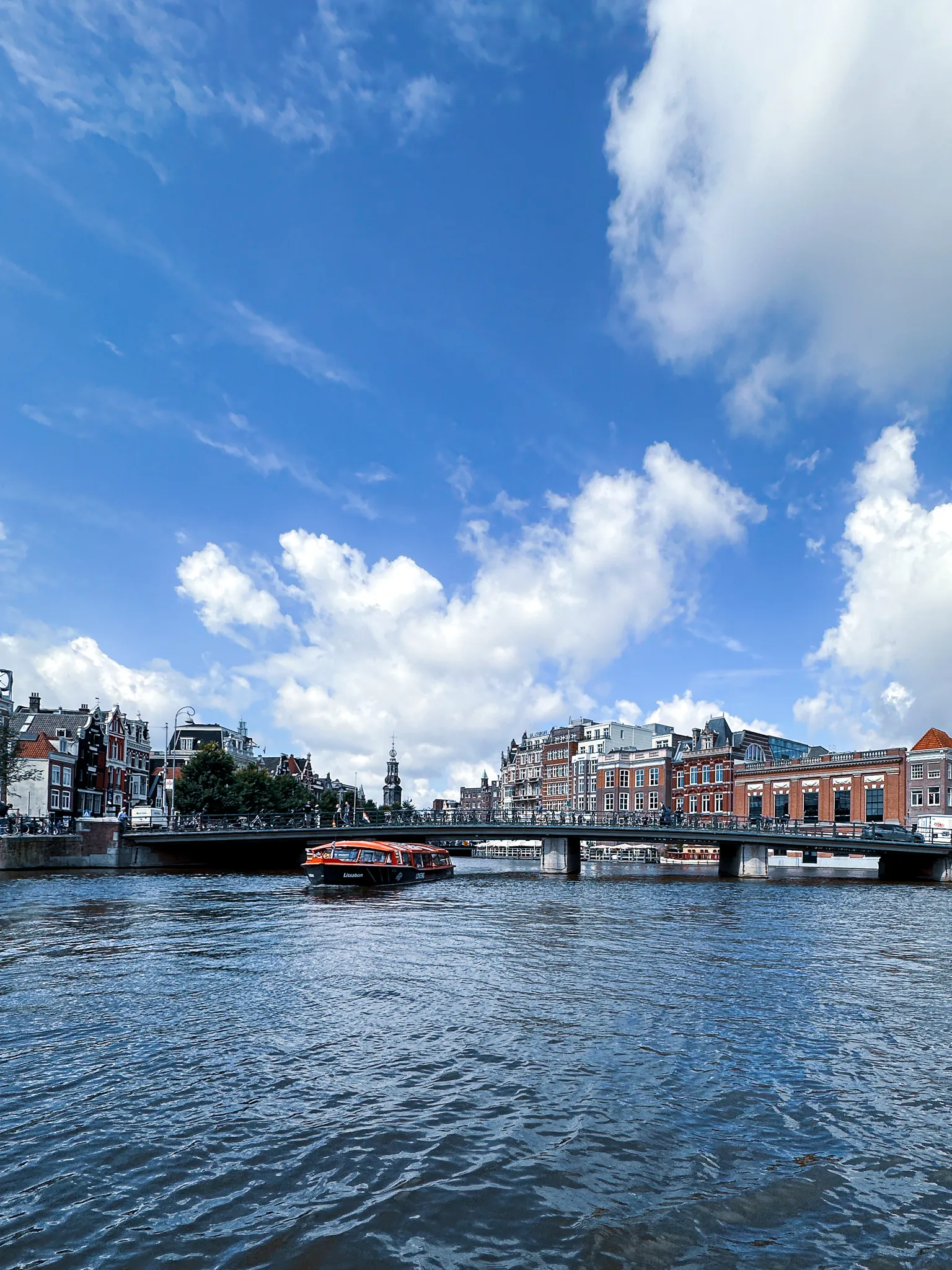 A low bridge lined with parked bicycles spans the wide grey water, an orange and black tour boat sliding under it and a slim spire beyond, three quarters of the frame given over to blue sky and cumulus