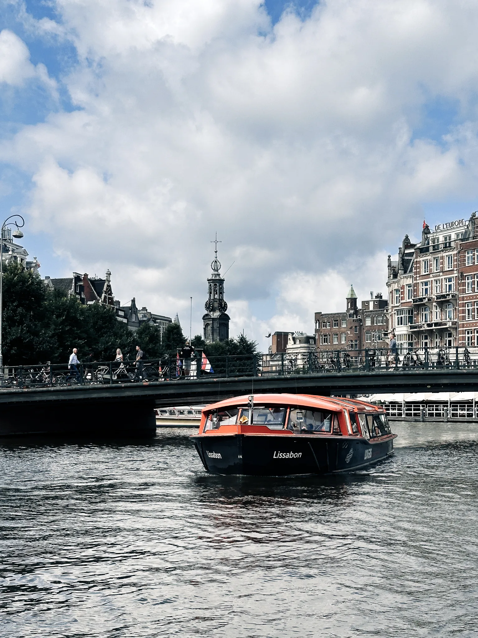 A canal tour boat named Lissabon noses out from under a bridge crowded with cyclists and walkers, the Munttoren's clock spire standing over the rooftops behind it