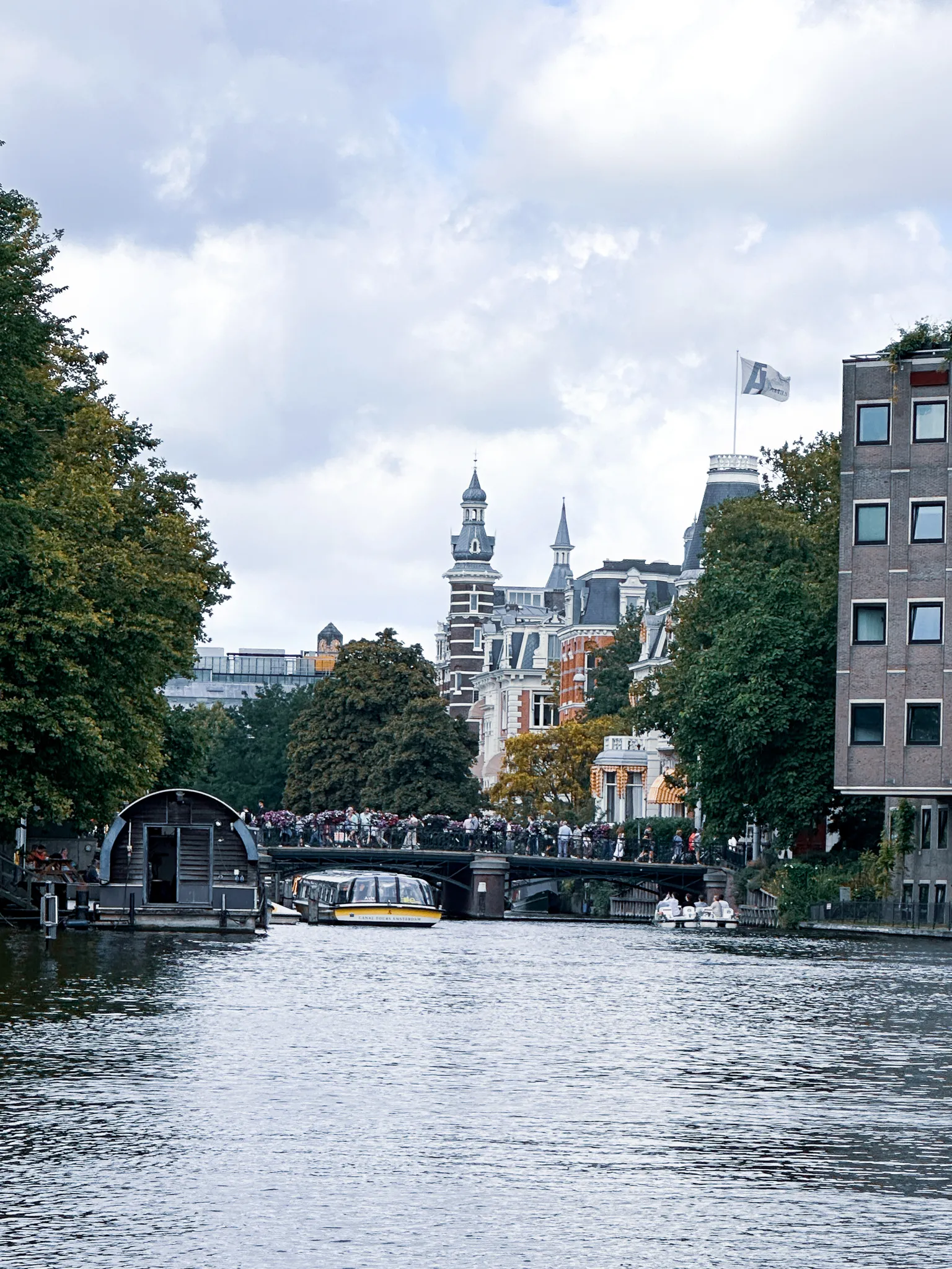 Looking up the canal toward a bridge packed shoulder to shoulder with people and bikes, a small yellow tour boat waiting beneath it and turrets and a flagpole rising over the trees beyond
