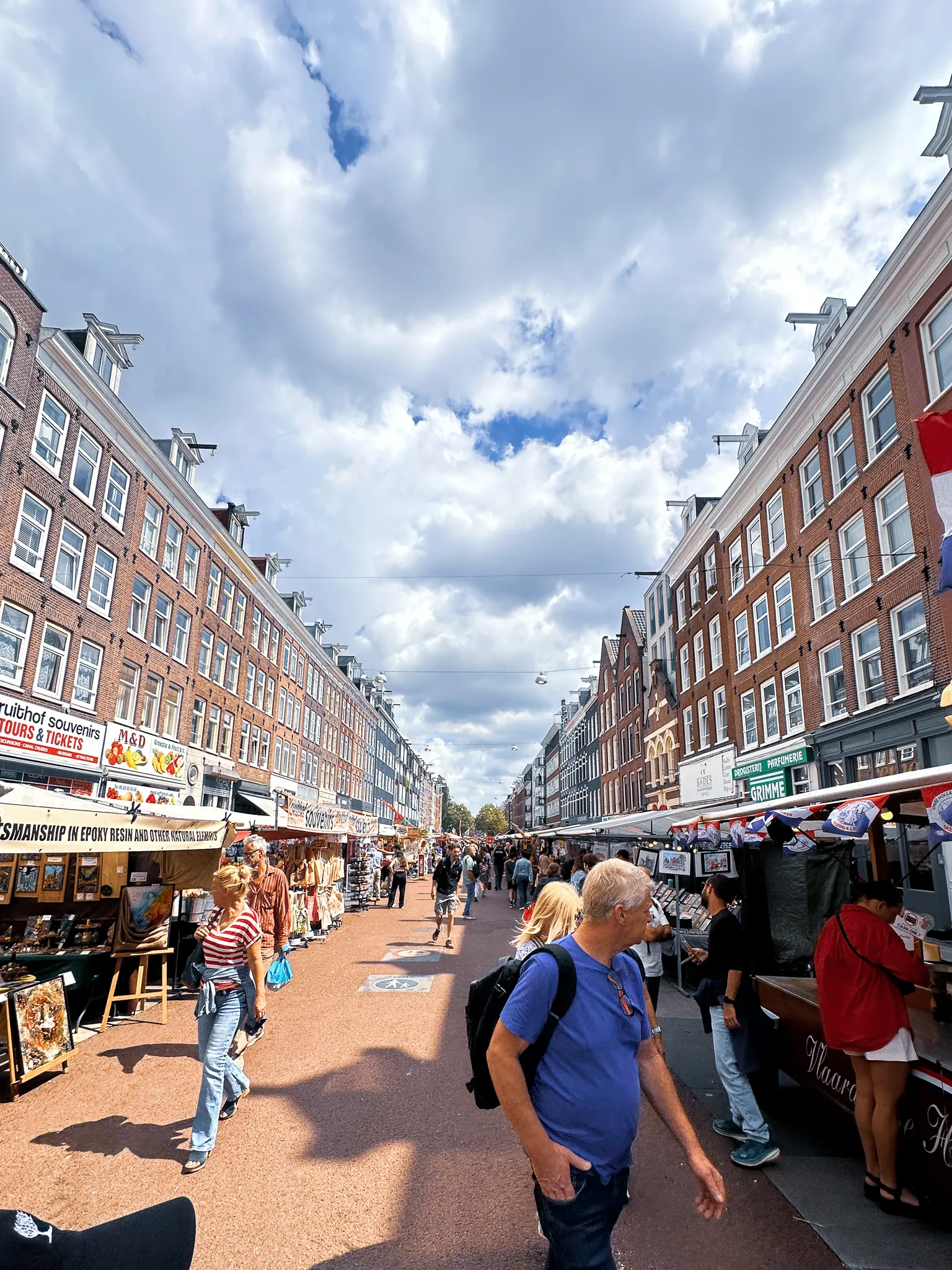The Albert Cuyp under big cloud, stalls and awnings down both sides of the long straight street beneath brick tenements, shoppers filling the middle and a sign for epoxy-resin craftwork on the left