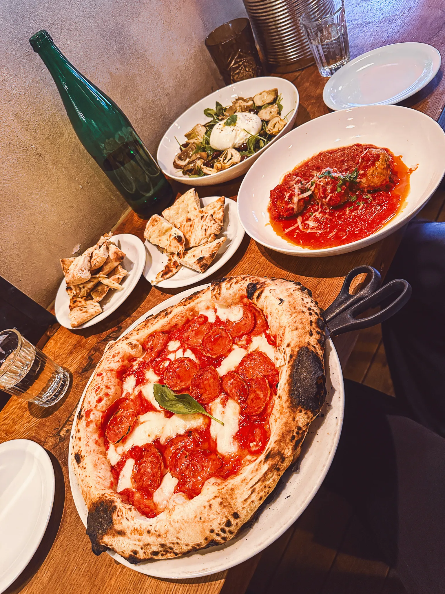 A wooden table shot from above - a blistered pepperoni pizza in its pan, meatballs under tomato and parmesan, burrata with mushrooms and artichokes, torn flatbread and a tall green bottle