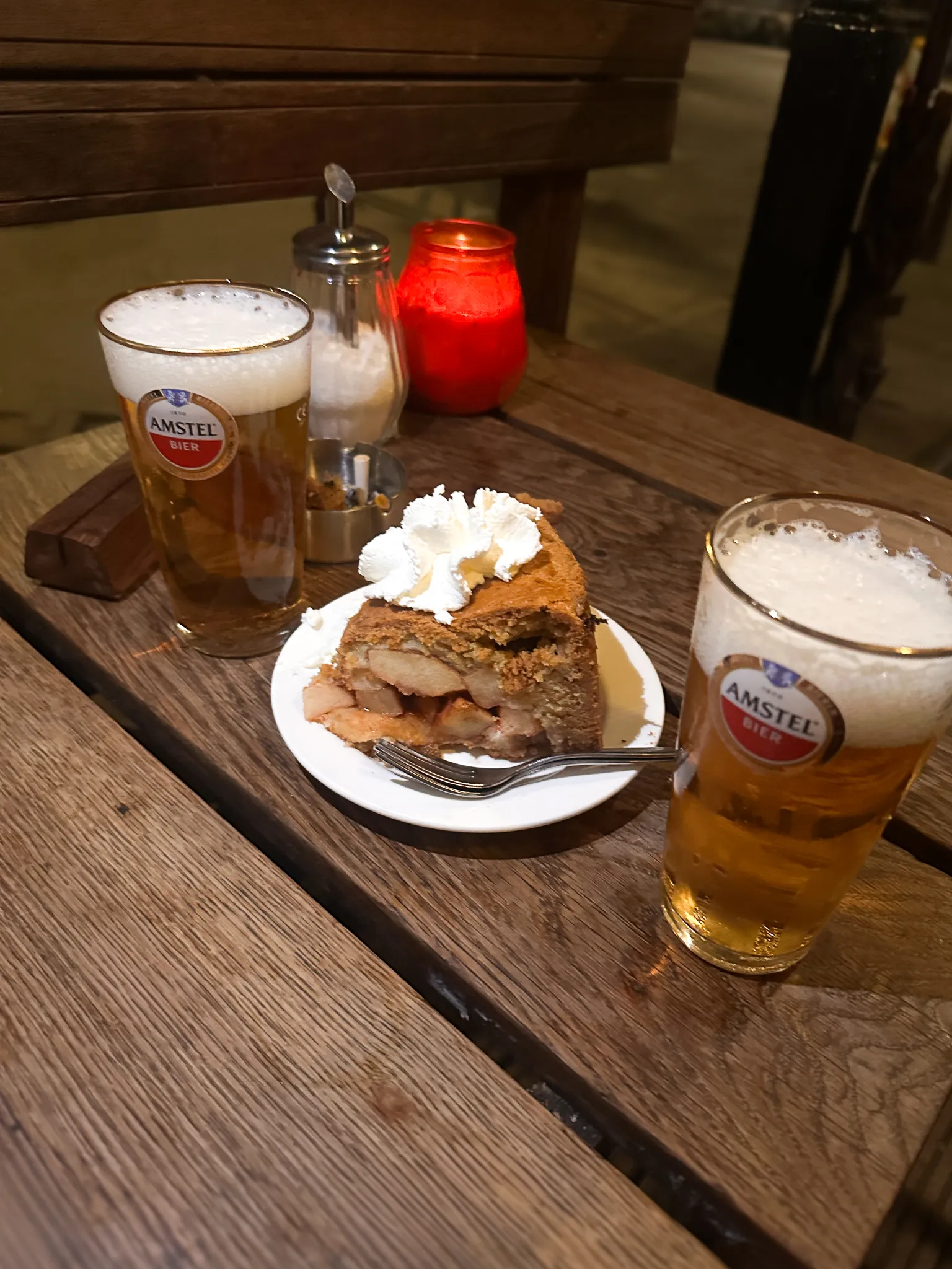 A wedge of Dutch apple pie under a heap of whipped cream sits between two glasses of Amstel on a scarred wooden table, a red candle and a sugar shaker behind