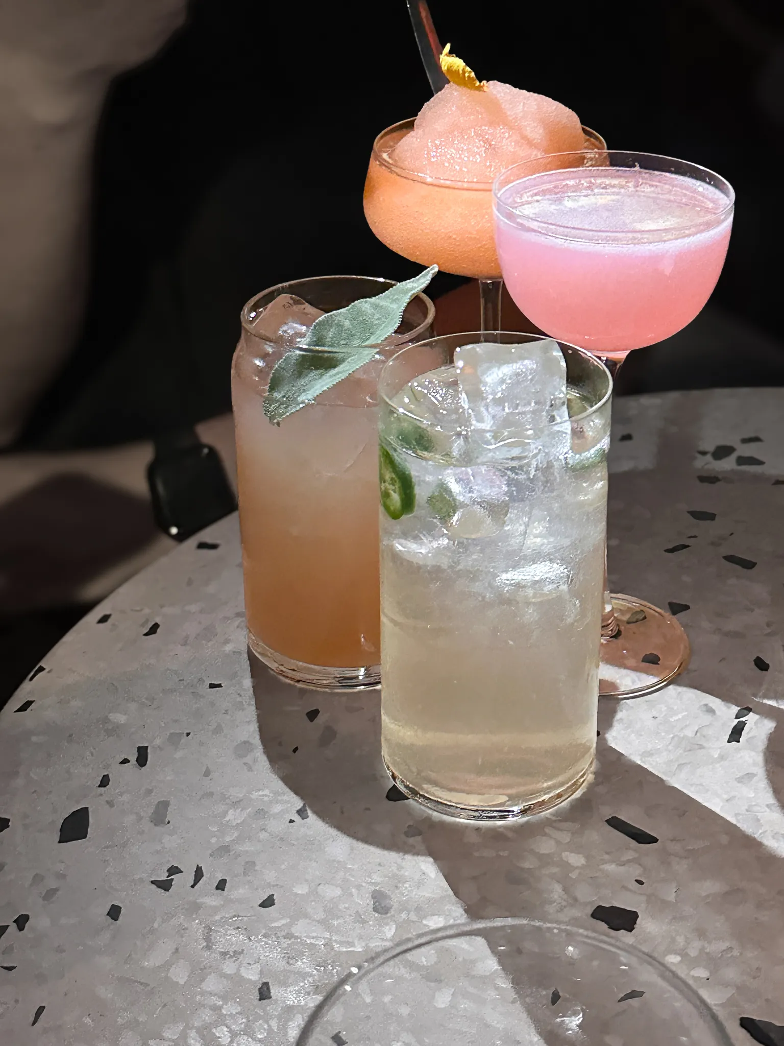 Four cocktails crowded on a speckled terrazzo table in hard low light - a coupe crowned with a grapefruit slice, a pink foamed sour, a sage-garnished highball and one packed with ice and jalapeño