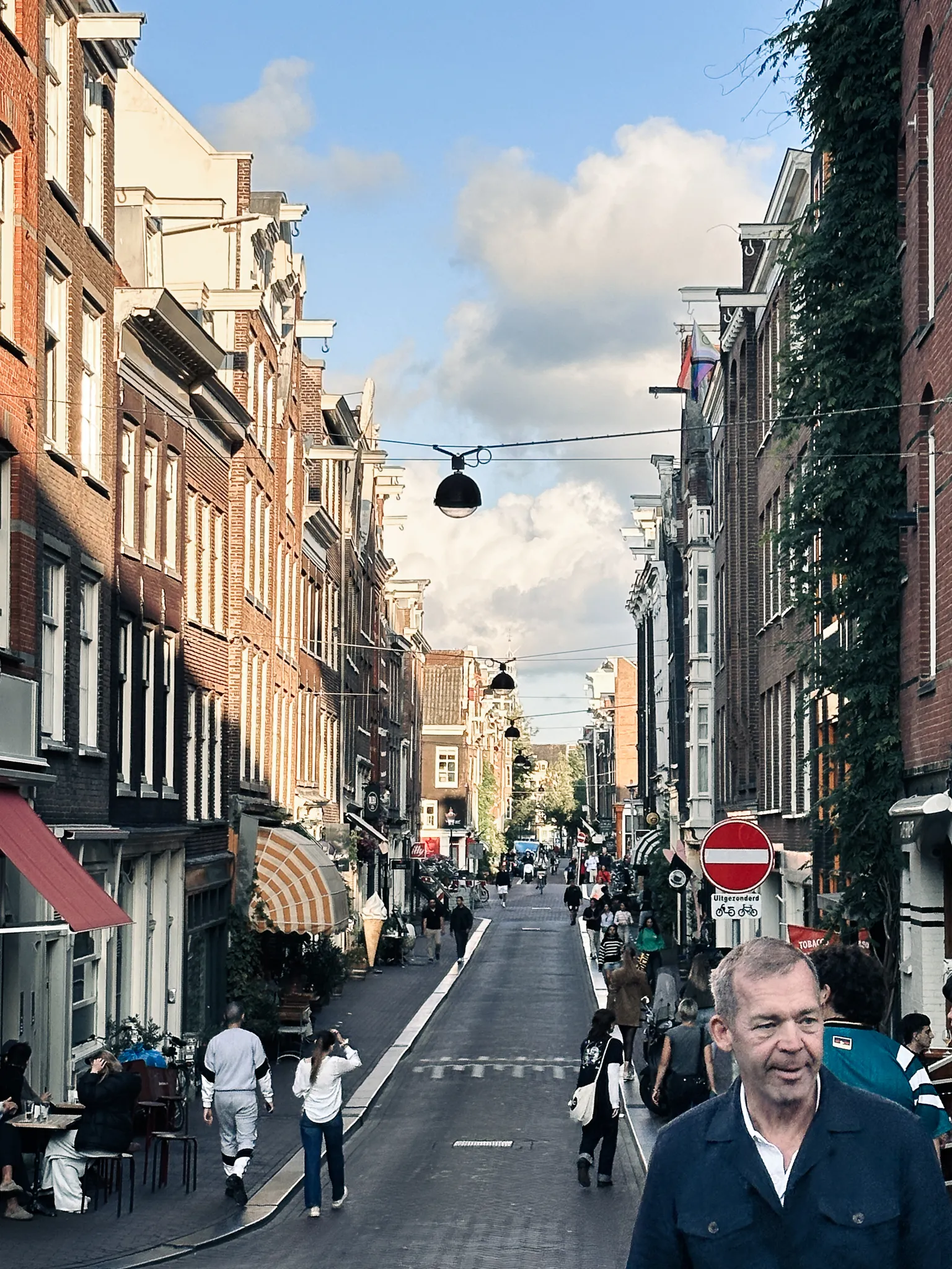 A narrow brick shopping street with a gelato sign and awnings down one side and ivy climbing the other, shoppers spread along the middle and a passer-by's face filling the bottom corner of the frame