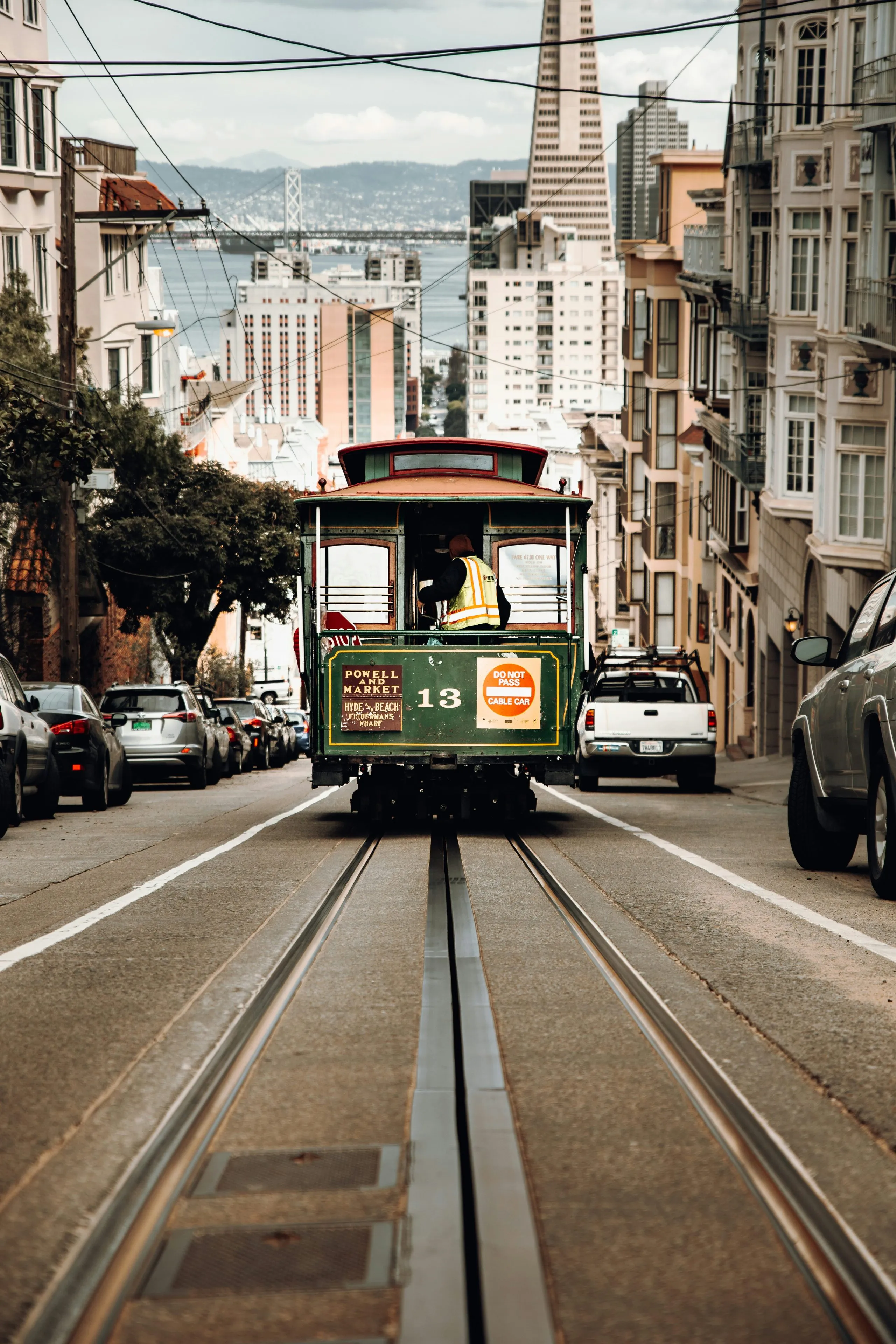 A green cable car head-on on its tracks, parked cars either side and the Transamerica Pyramid rising behind