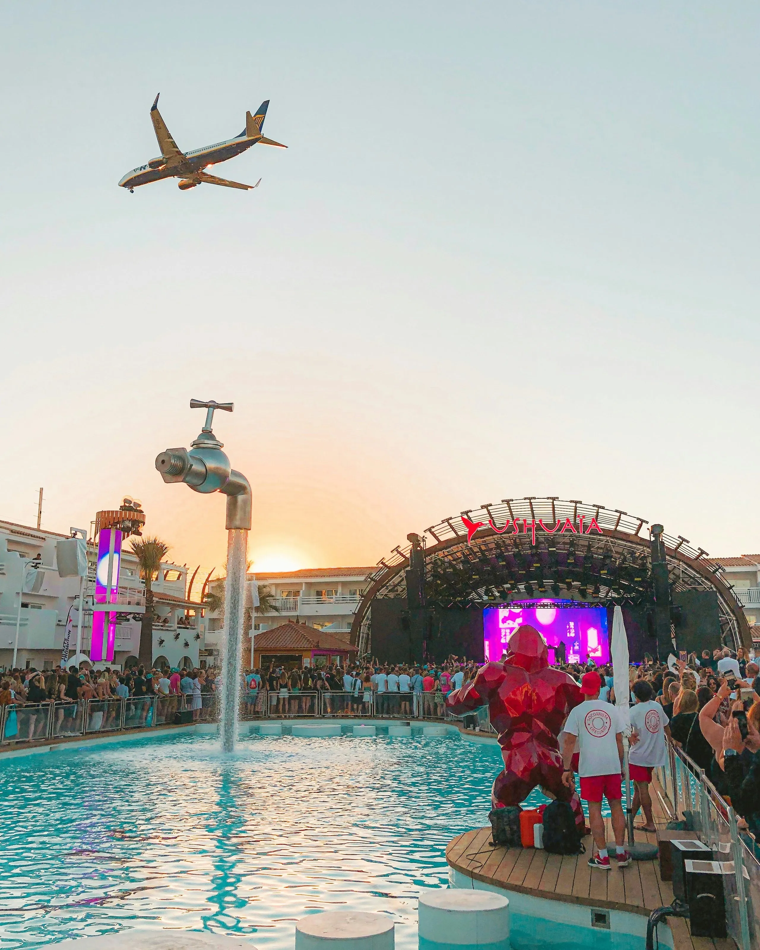 A poolside stage at sunset with a plane crossing the sky above, a figure in red on the deck and a foam cannon running