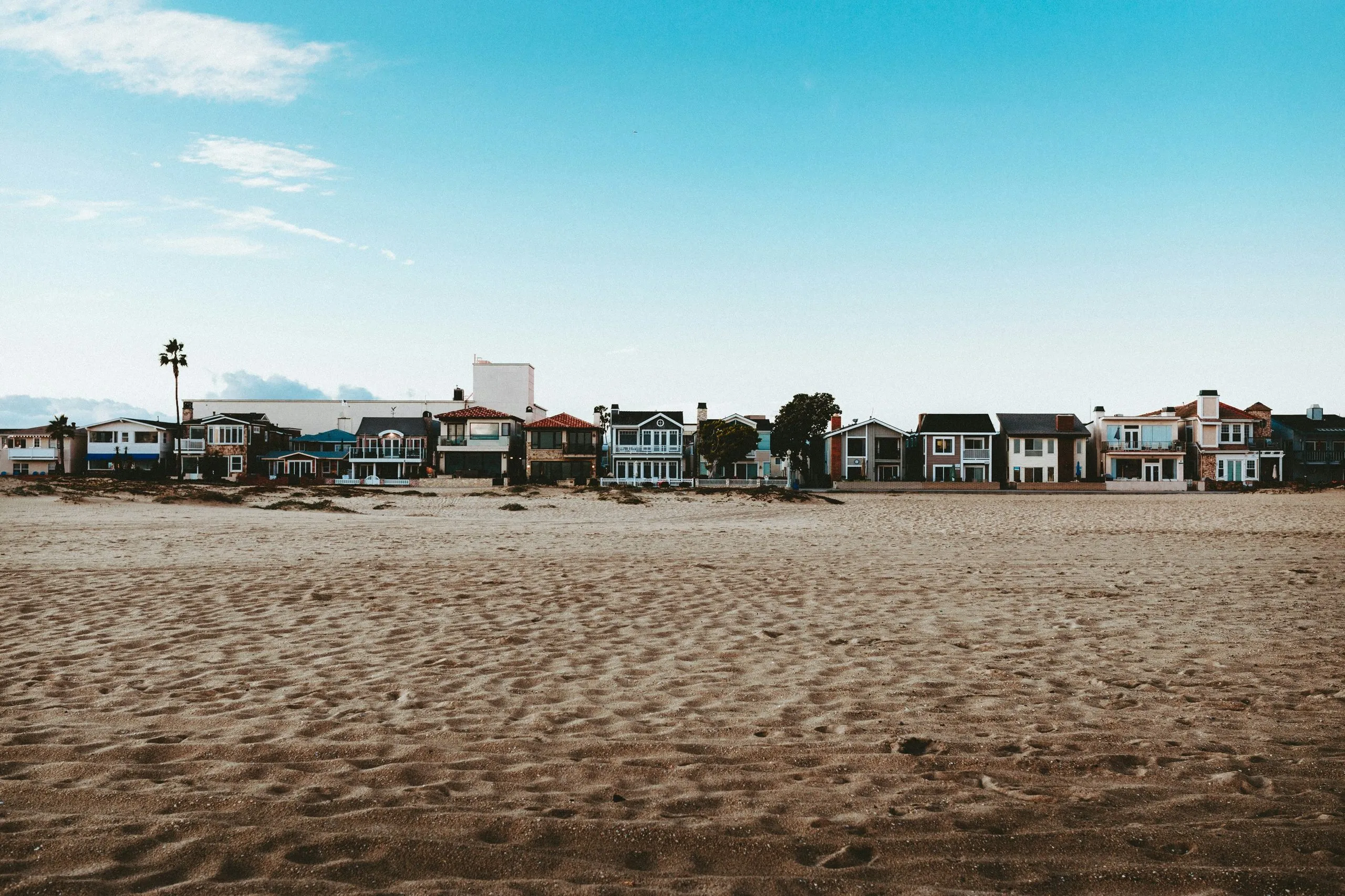 A wide stretch of empty rippled sand with a row of beach houses along the far edge under a bright sky