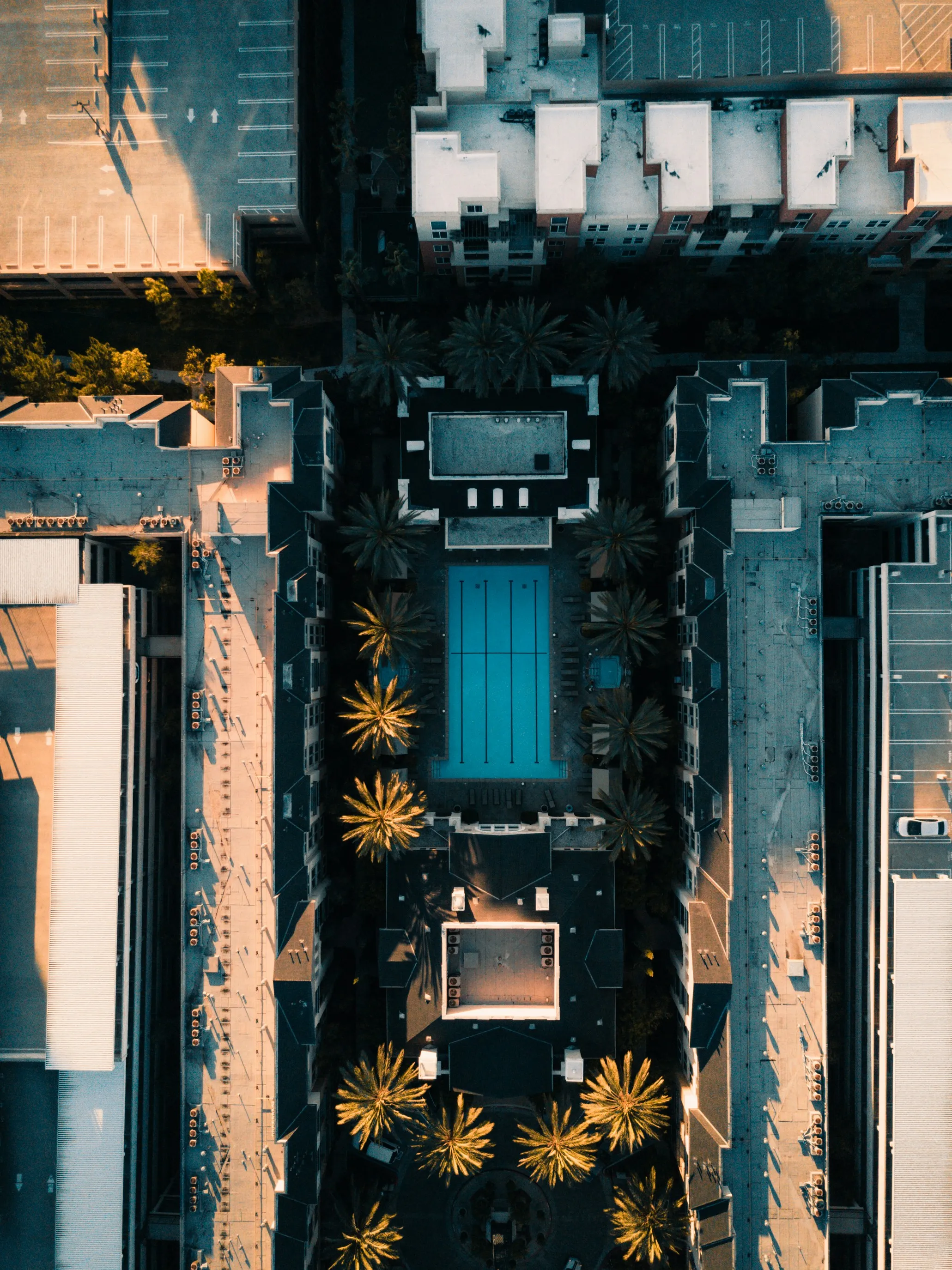 Looking straight down on a resort pool, its turquoise water set between white buildings and rows of palms