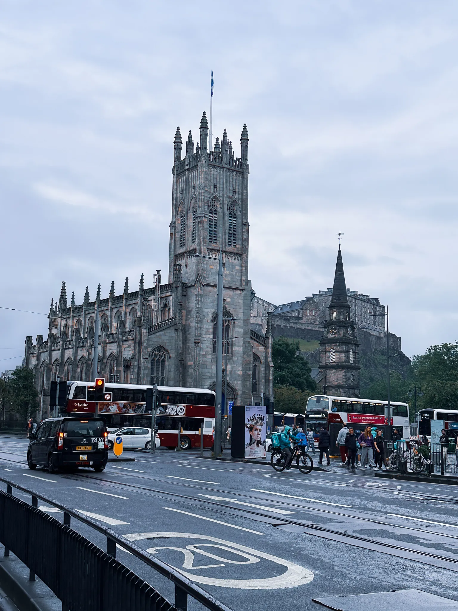The tower of St John's at the West End with a saltire flying, St Cuthbert's spire and the castle stacked behind, buses held at the lights