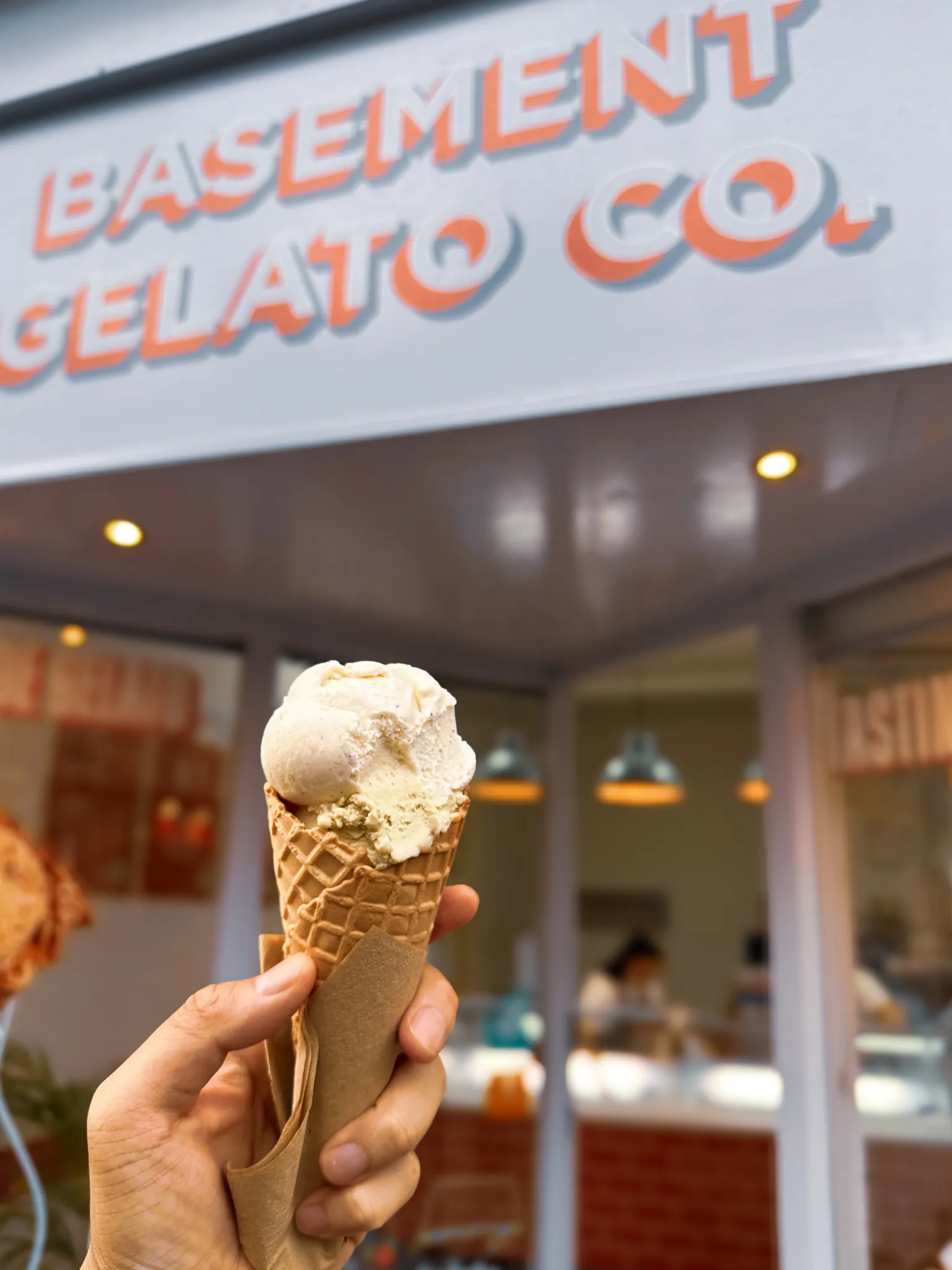 A waffle cone held up outside Basement Gelato Co., one pale scoop over pistachio, the shop sign going soft behind it