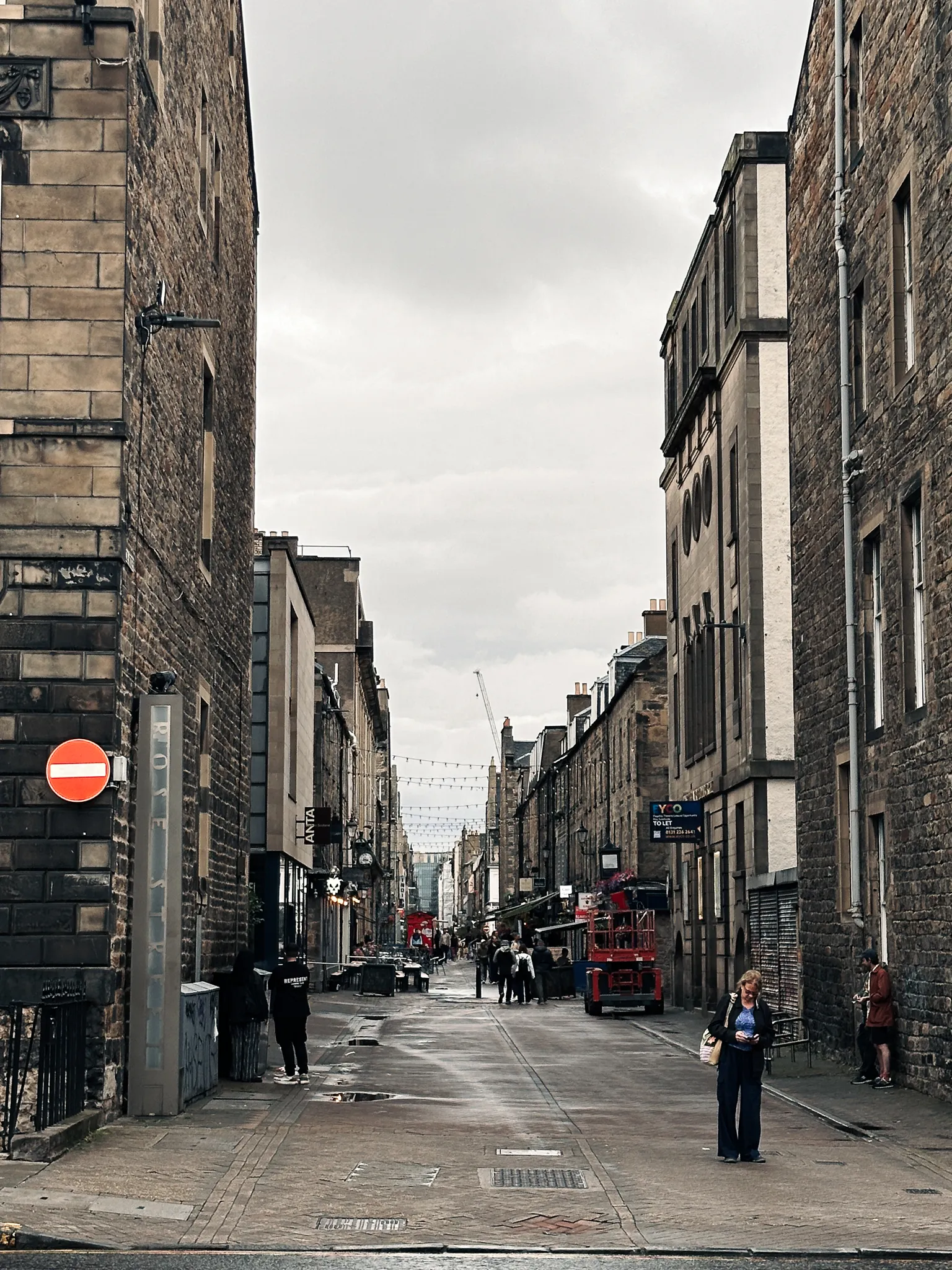 Rose Street emptying out in the evening - string lights overhead, puddles between the setts and shop signs lit down the lane