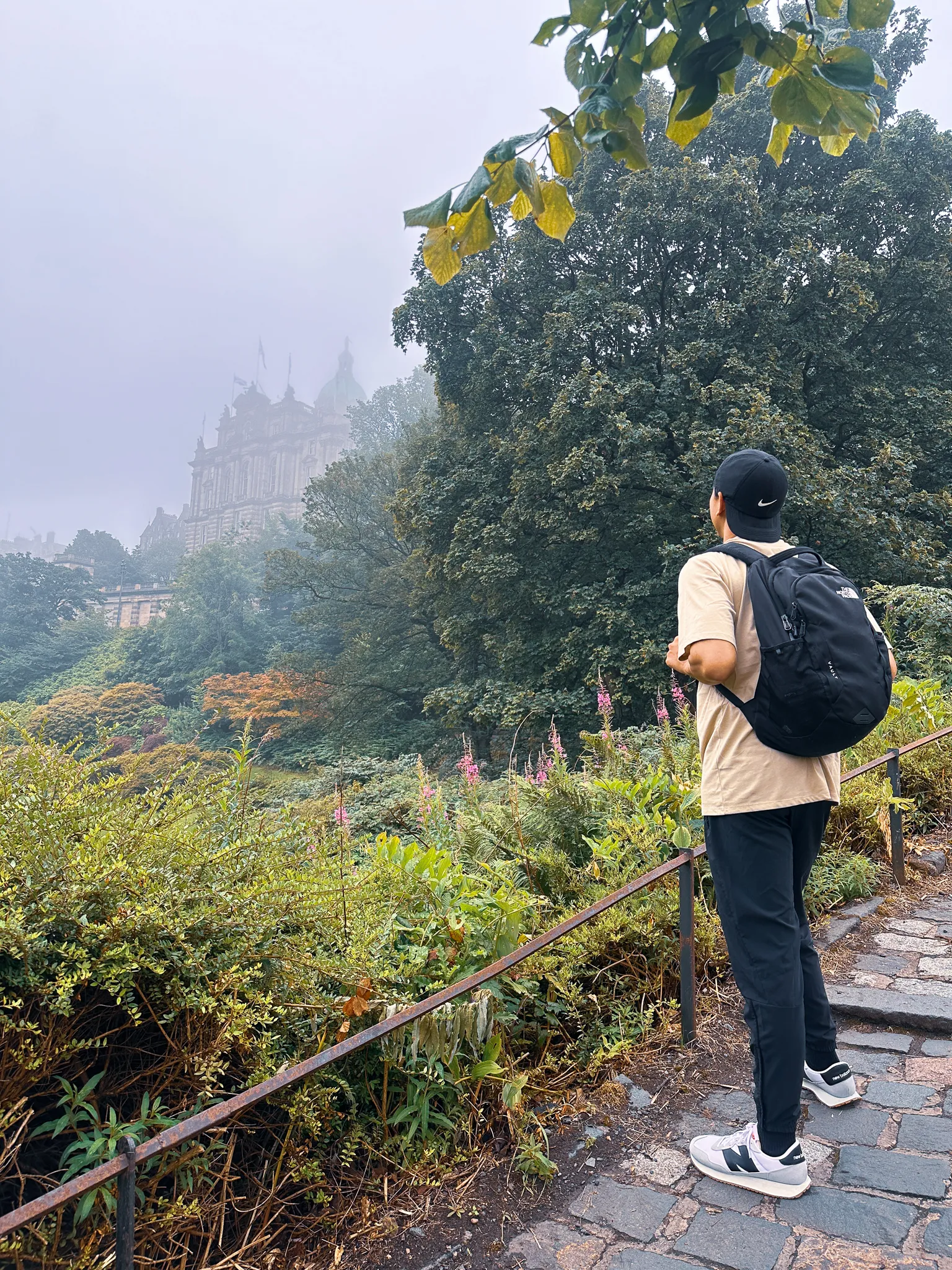 Stopped on the cobbled path above the gardens, cap and backpack on, watching the bank's domes come and go in the haar