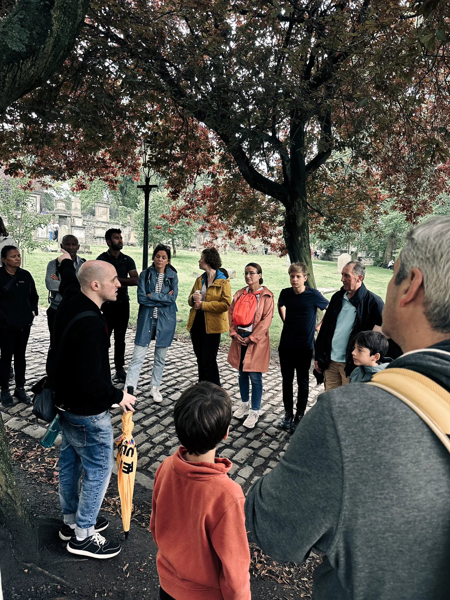 A walking tour ringed under the copper beech in Greyfriars Kirkyard, the guide mid-story with a furled yellow umbrella, headstones on the lawn behind