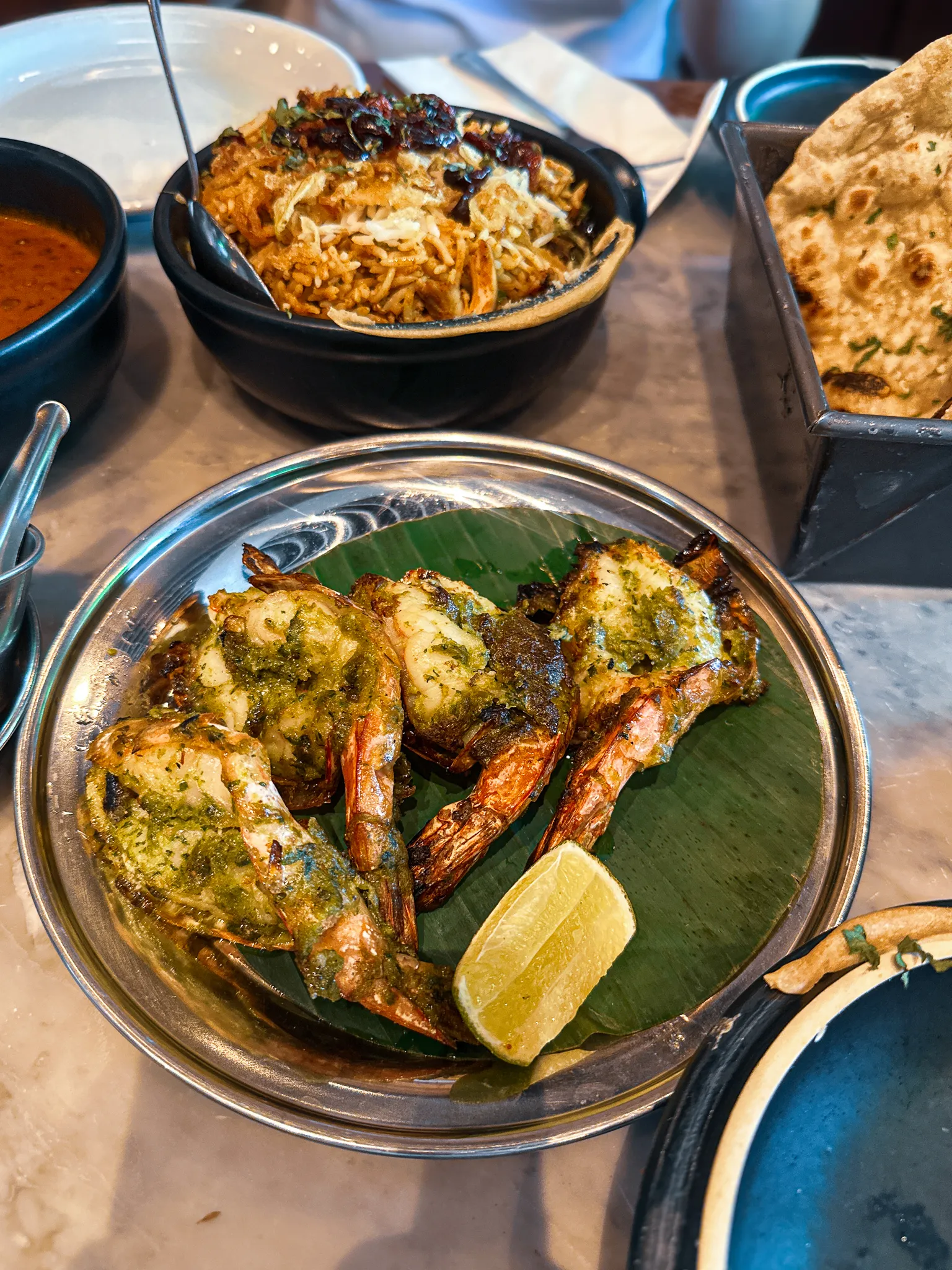 Tandoori king prawns green with herbs on a banana leaf with a lime wedge, biryani in a black pot and roti standing in a tin behind