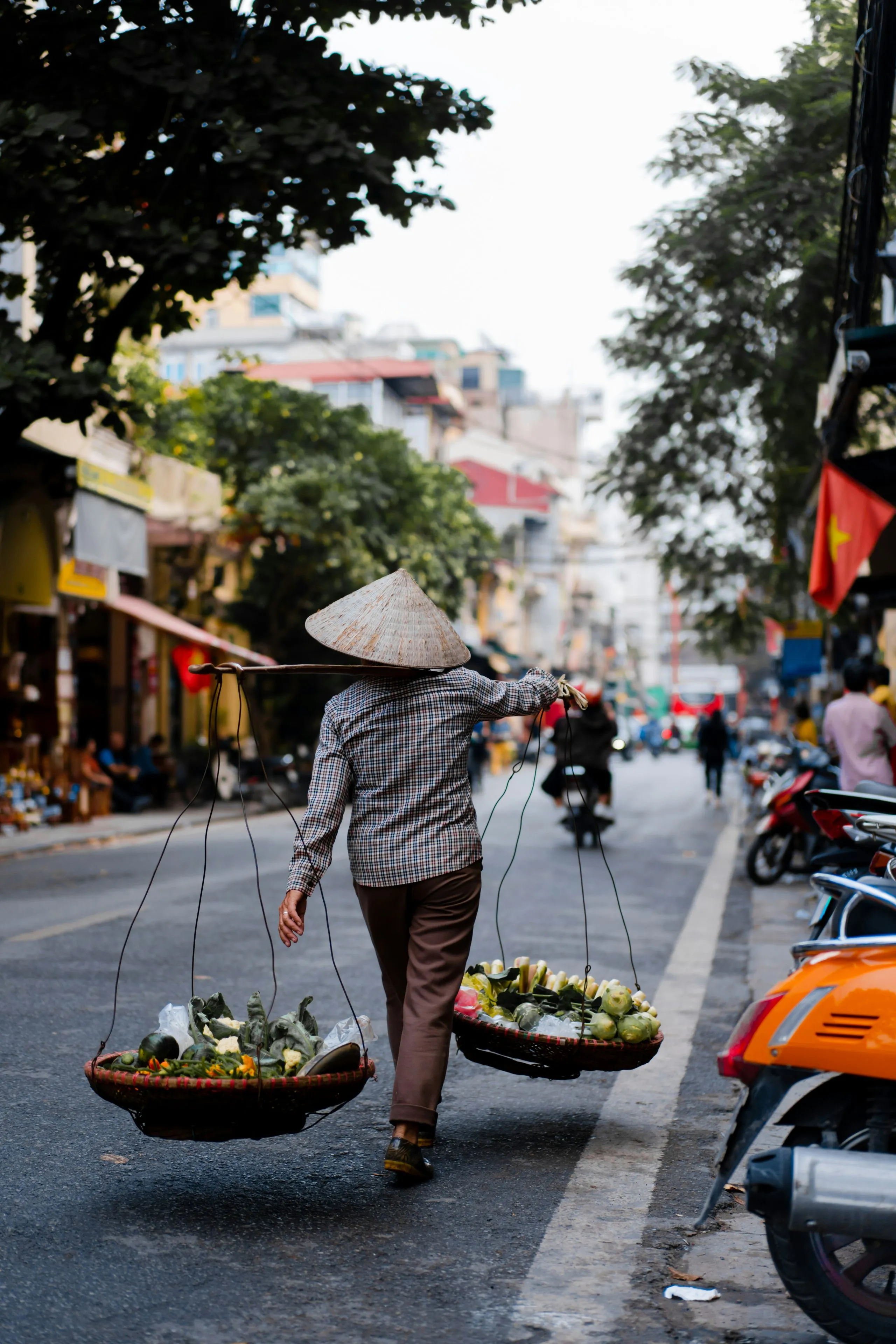 A seller in a conical hat walks down the middle of a shopfront street carrying two shallow baskets of flowers on a shoulder pole