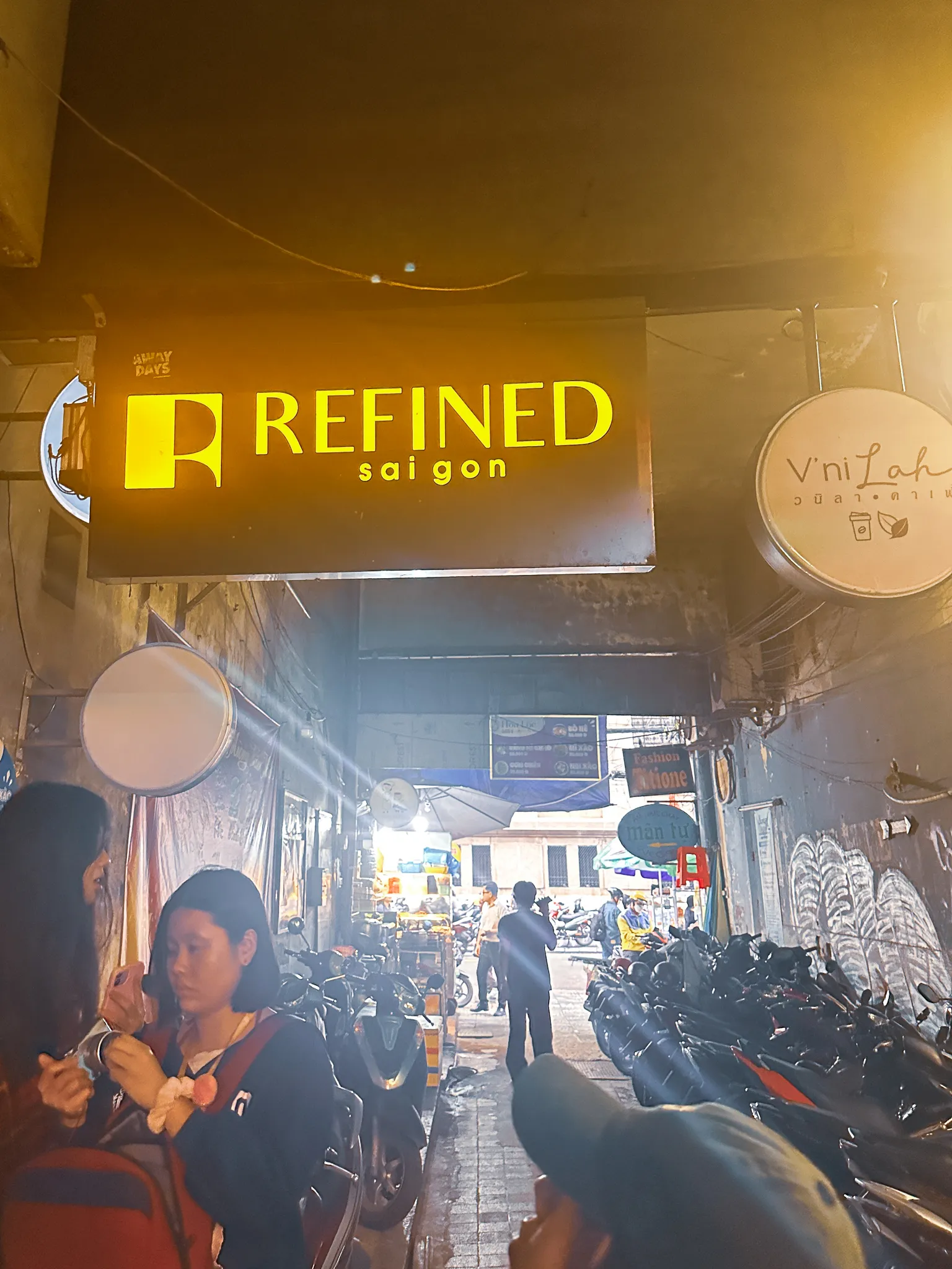 A glowing yellow sign for Refined Saigon over a narrow alley of parked scooters, cafe signs and food stalls