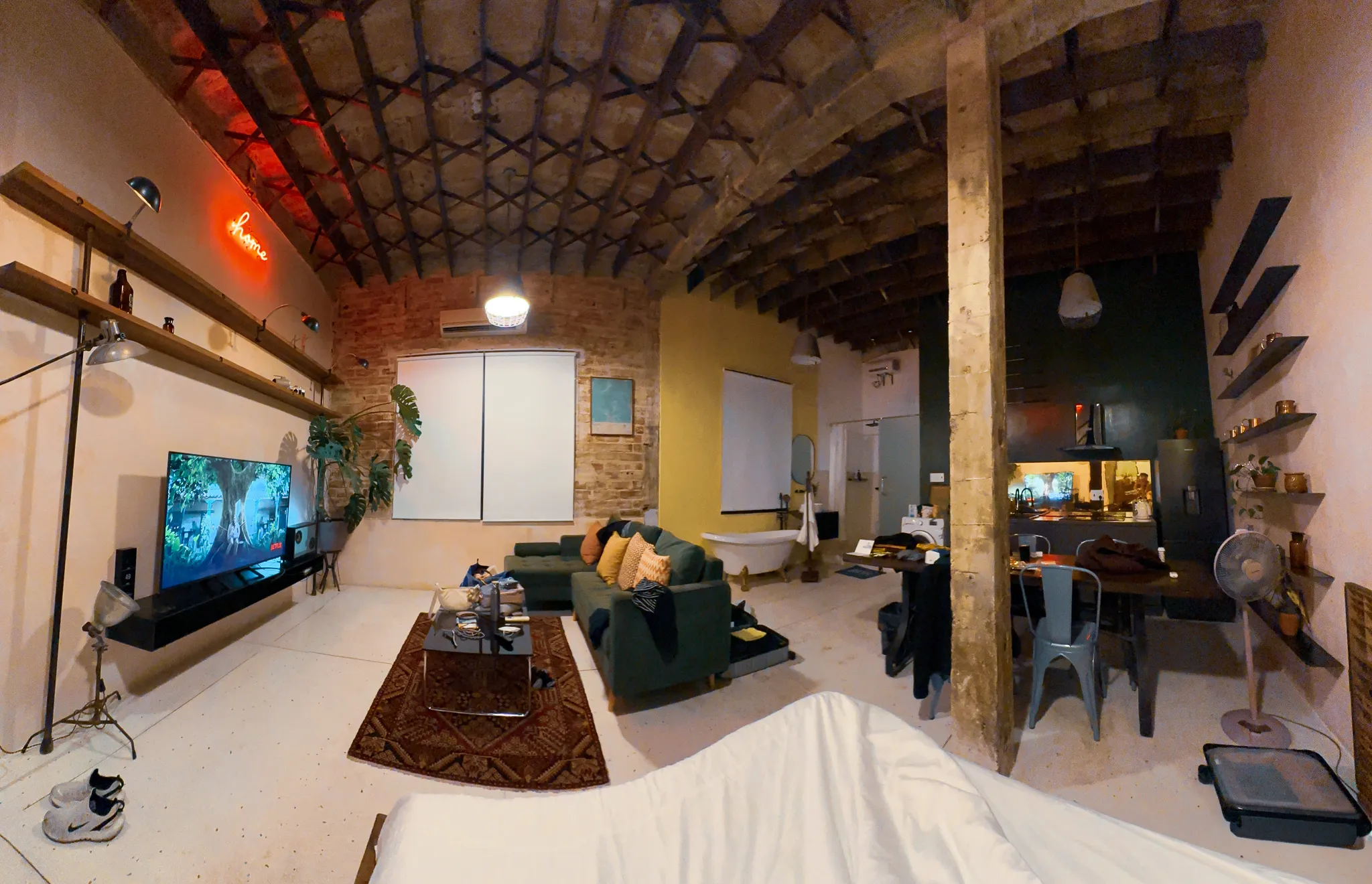 A brick-vaulted loft with a red neon home sign, green sofa, clawfoot bathtub and kitchen sharing one open room