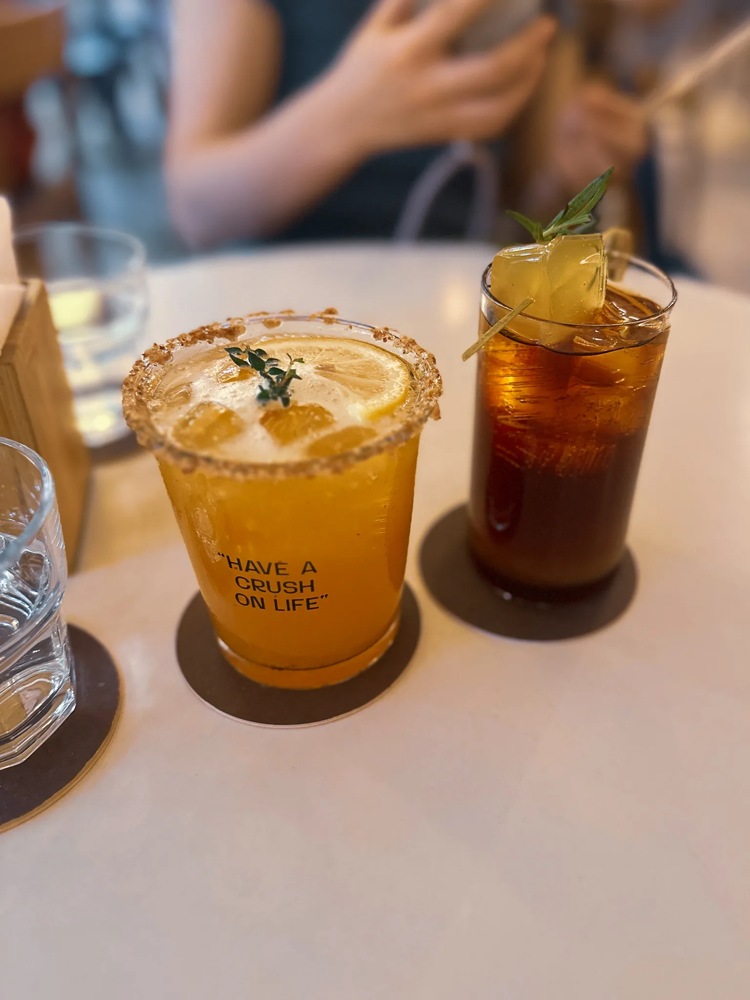 An orange drink with a sugared rim printed have a crush on life, beside a tall cold brew with an ice skewer