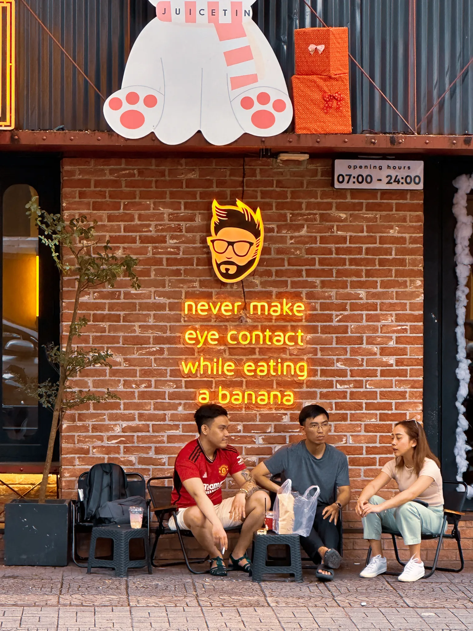 Three friends on low chairs outside a juice bar, under a neon sign reading never make eye contact while eating a banana