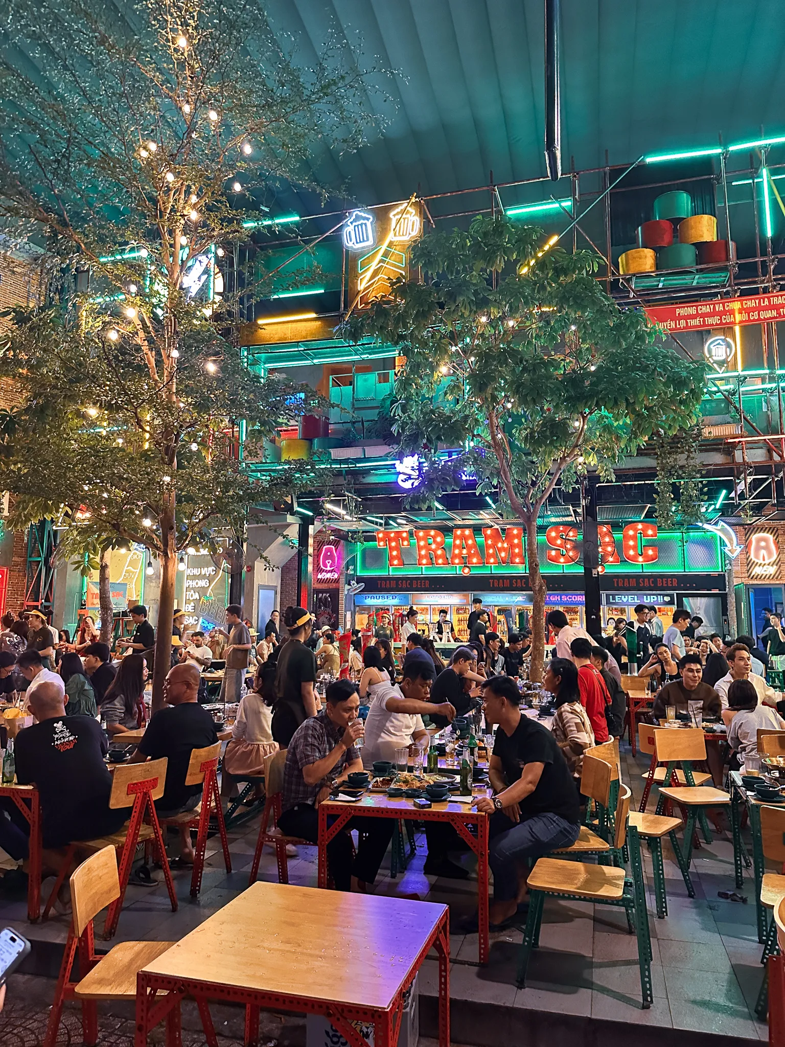 An open-air beer hall strung with lights, marquee letters spelling Tram Sac over trees and rows of full wooden tables