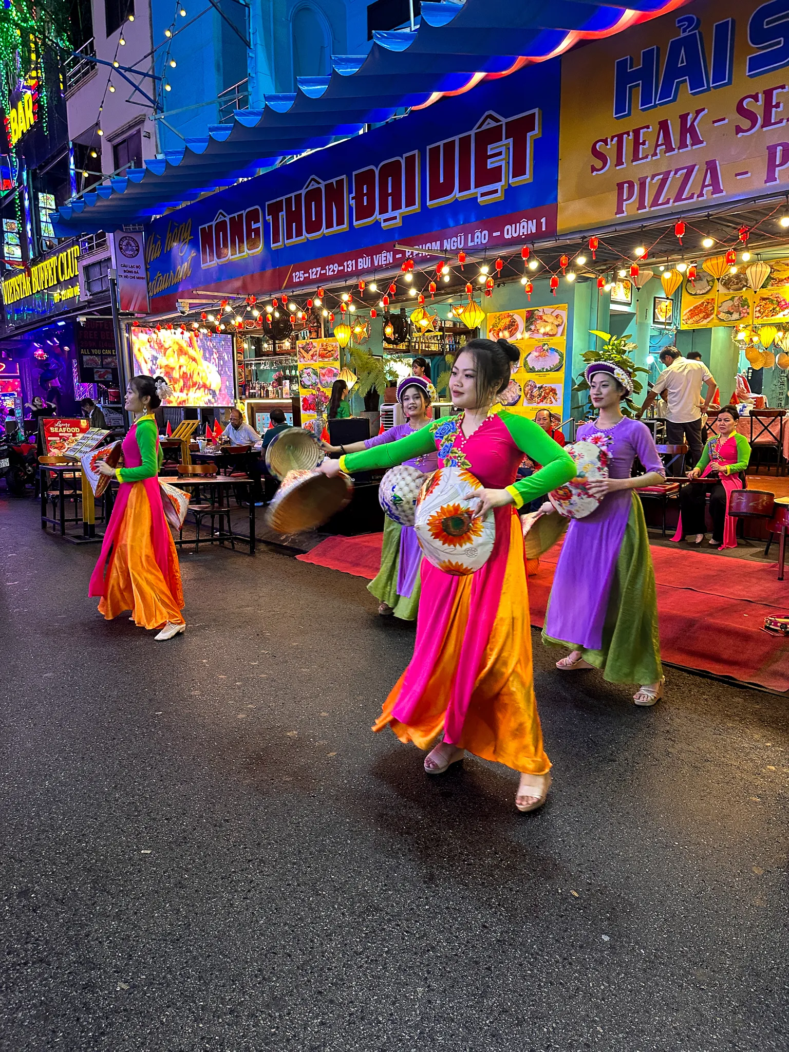 Dancers in bright ao dai spinning painted conical hats on Bui Vien, lanterns and menu boards lit behind them