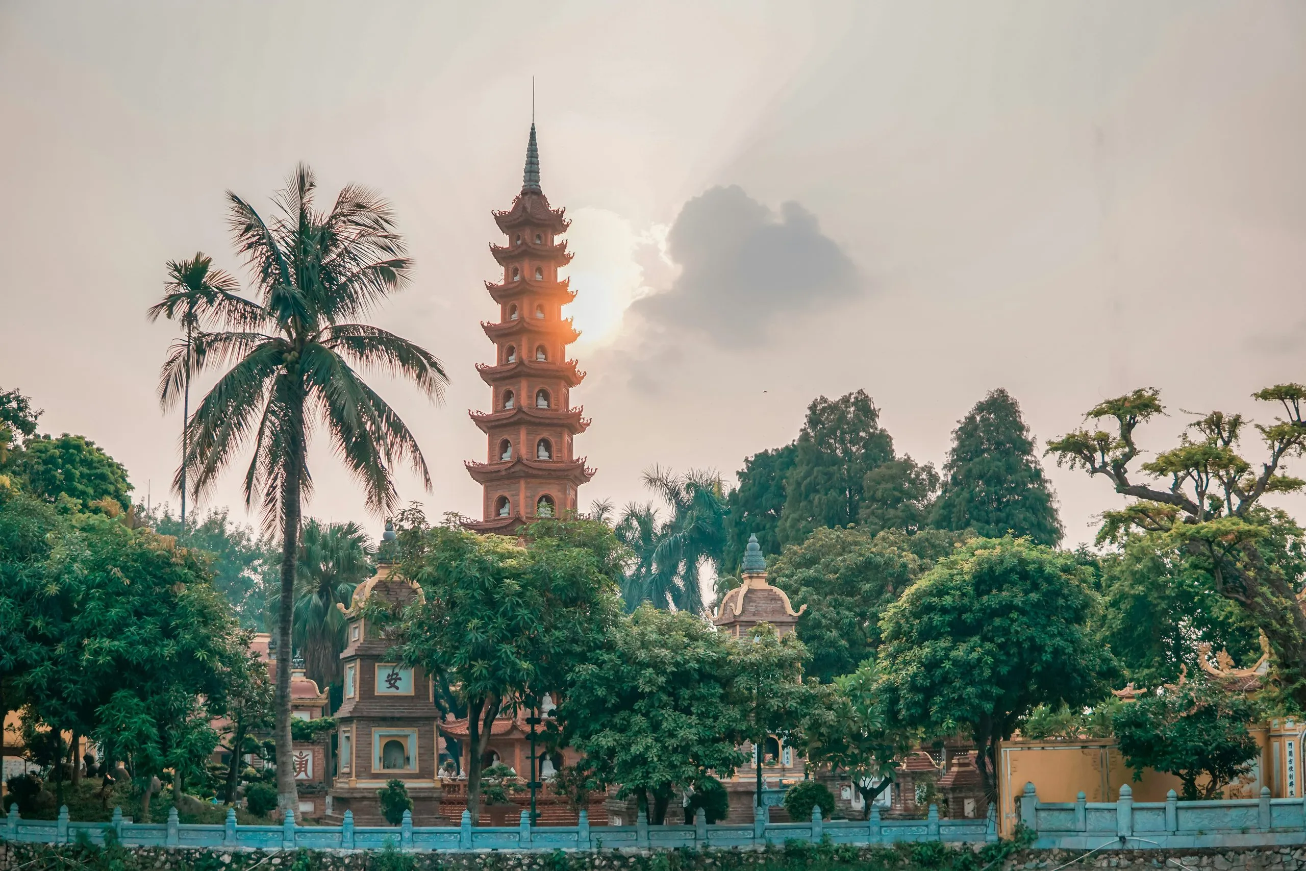 The tiered brick tower of Tran Quoc Pagoda rising above trees and palms against a pale, cloud-streaked evening sky