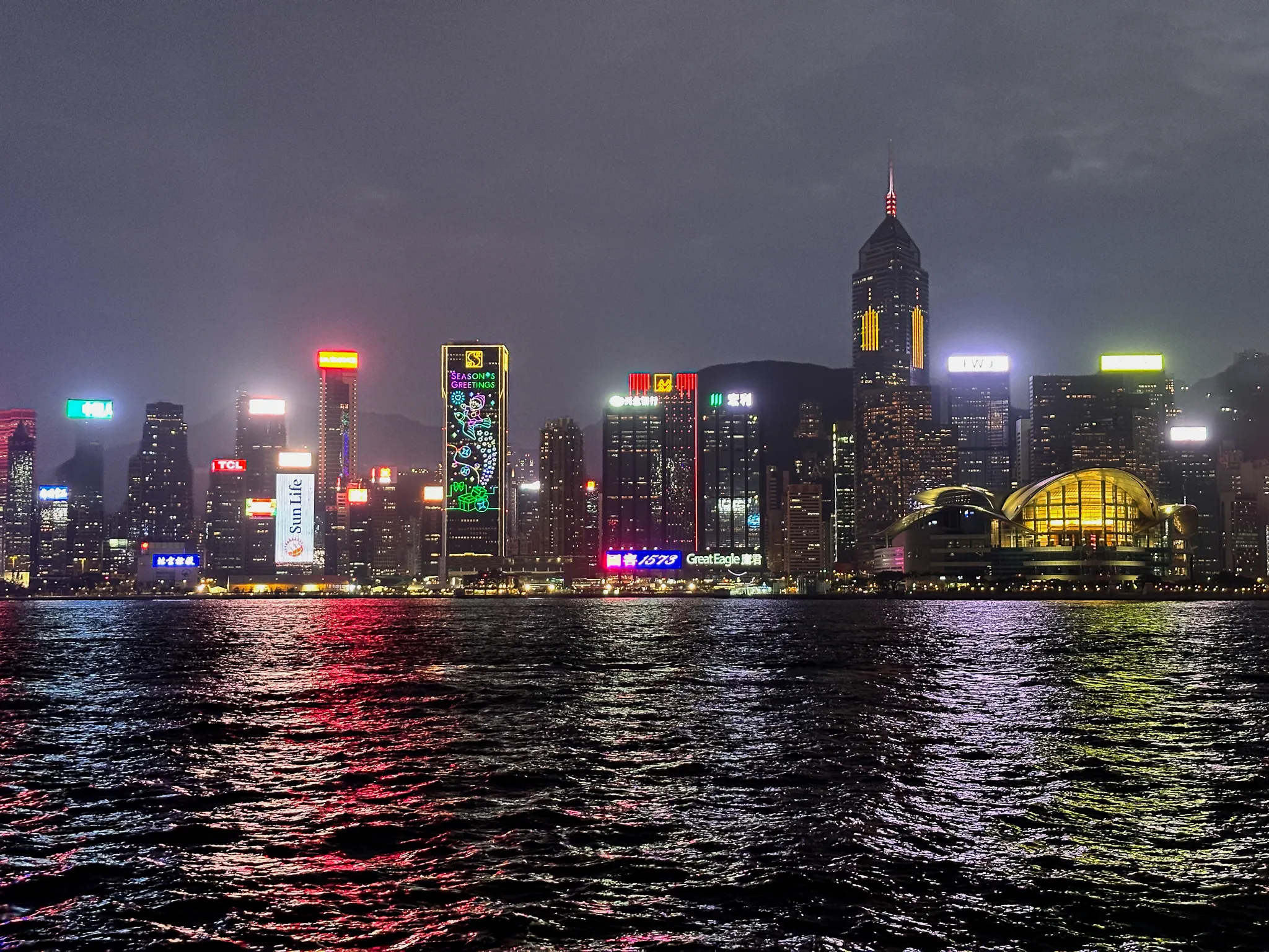 Victoria Harbour at night from the Kowloon side, the Wan Chai skyline lit end to end with a Season's Greetings display running down one tower and the convention centre's curved roof glowing gold, colour dragged across the water