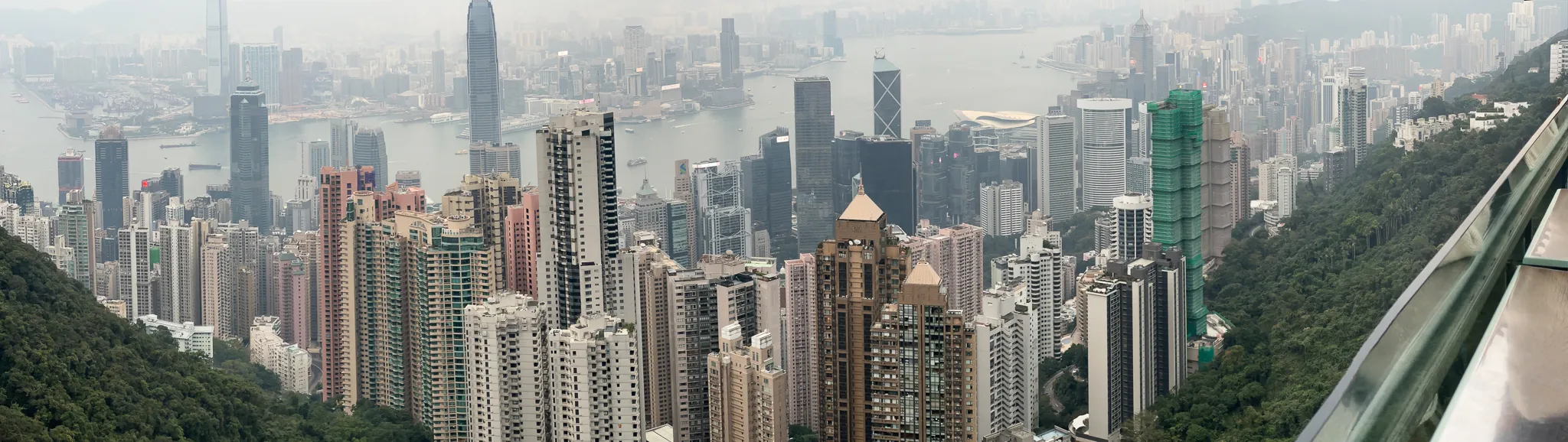 A wide view from Victoria Peak in daylight haze, towers rising straight out of the green hillside with the harbour flat behind them and the viewing deck's metal rail curving in at the right