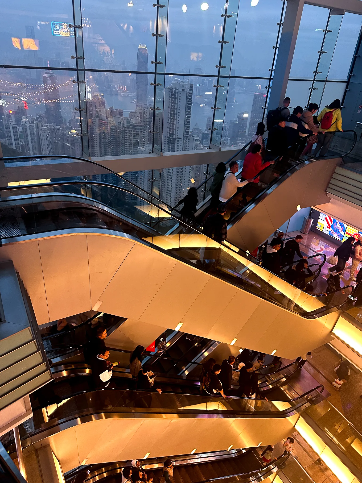 The same crossing escalators framed tighter, the glass wall and the dusk city filling the top of the frame