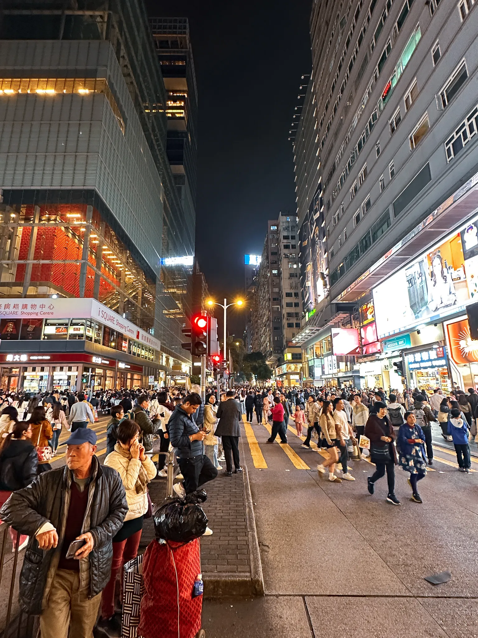 Nathan Road at night, a crowd held at a red light under stacked shop signs with the yellow crossing stripes running away into the traffic
