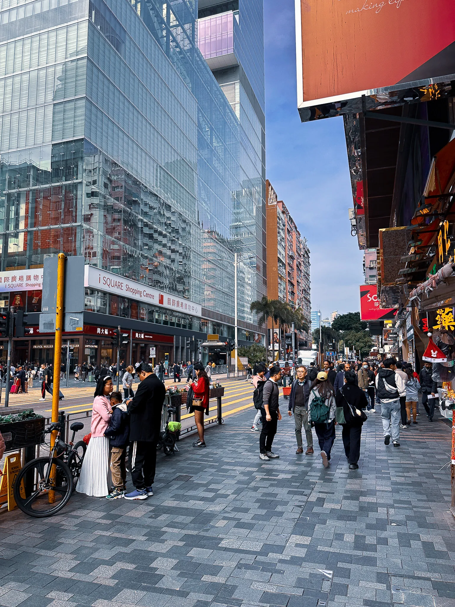 The same corner in winter daylight, iSQUARE's glass front on one side and a family stopped by a bicycle on the pavement, the street busy under a hard blue sky
