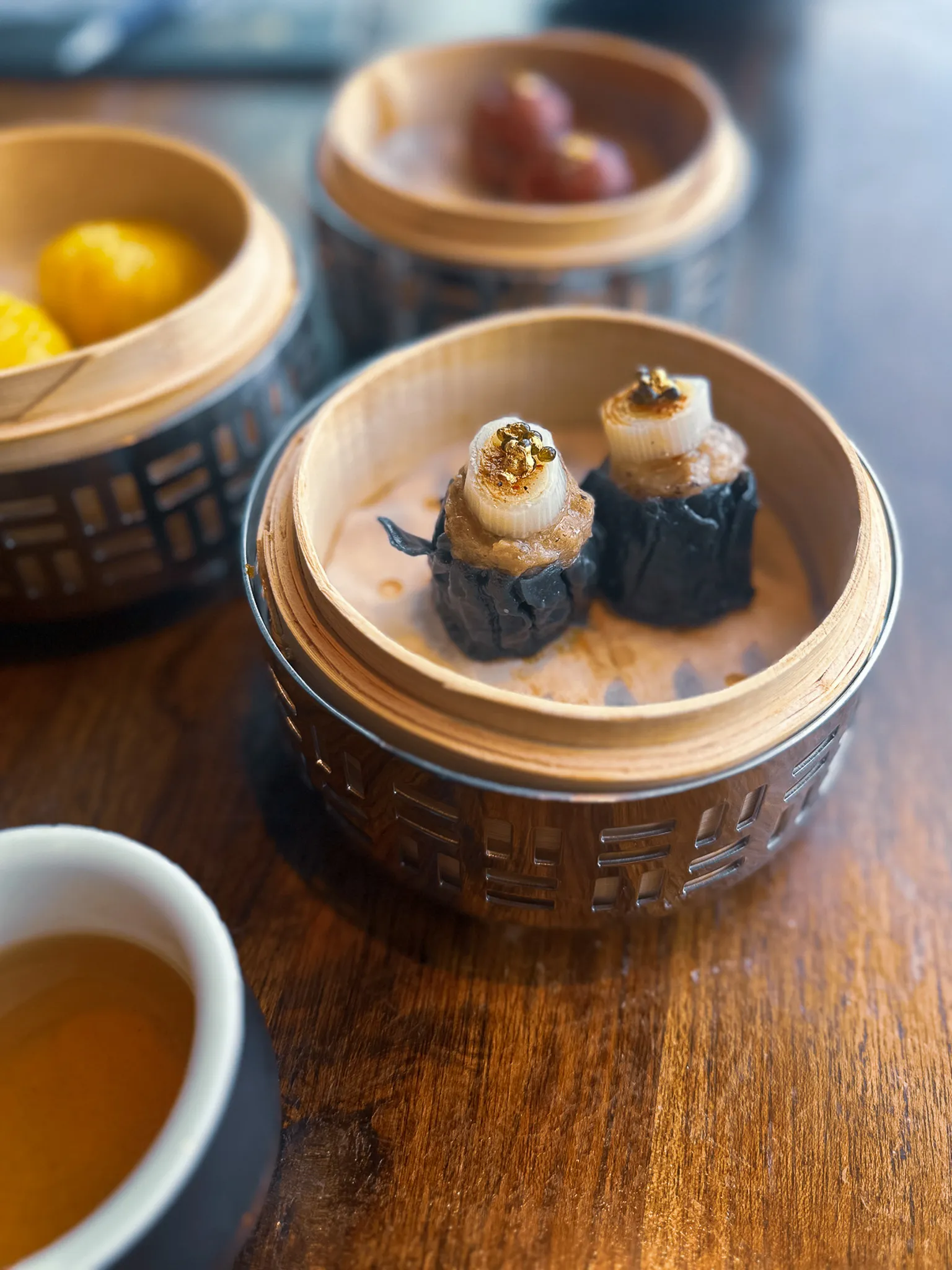 Three bamboo steamers in carved metal collars on a dark wood table, two charcoal-black parcels topped with leek and golden beads in the nearest one and a cup of tea at the edge of the frame
