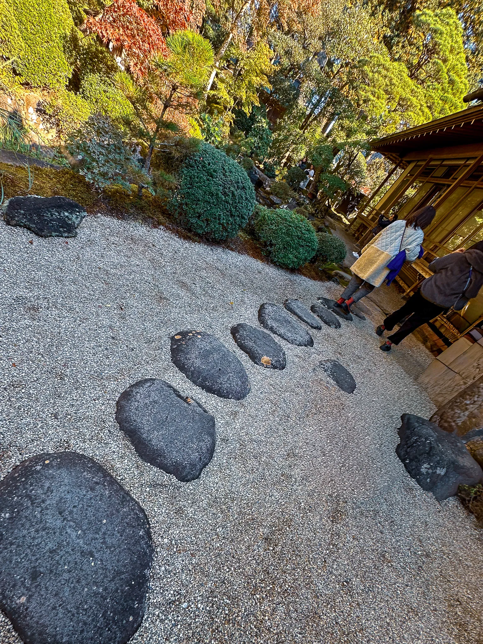Large flat lava stones set in a curve across a bed of raked white gravel, moss and clipped shrubs along the top edge, visitors on a wooden veranda to the right