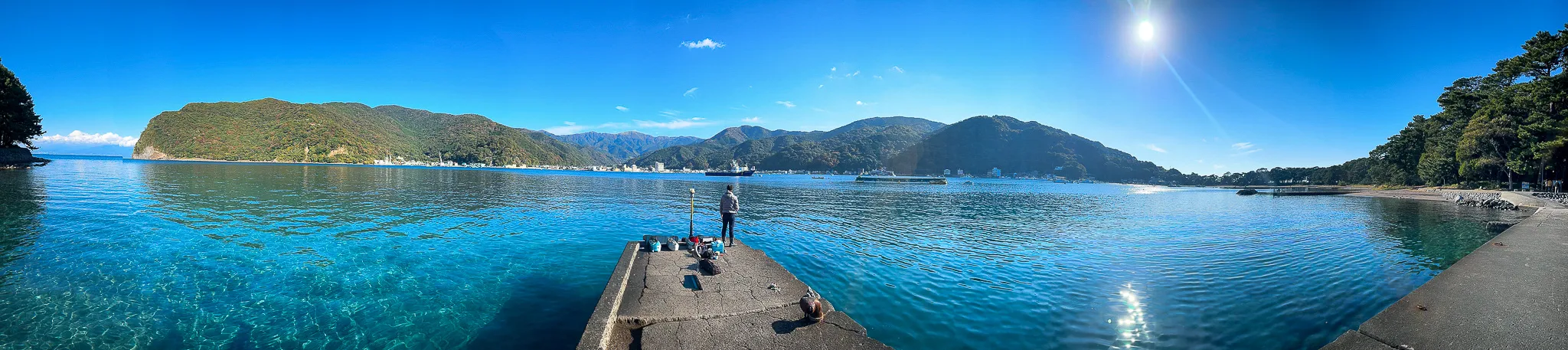 A very wide panorama of a bay so clear the bottom shows through, a concrete jetty running out to one figure fishing at the end, wooded headlands all around and the sun flaring on the right