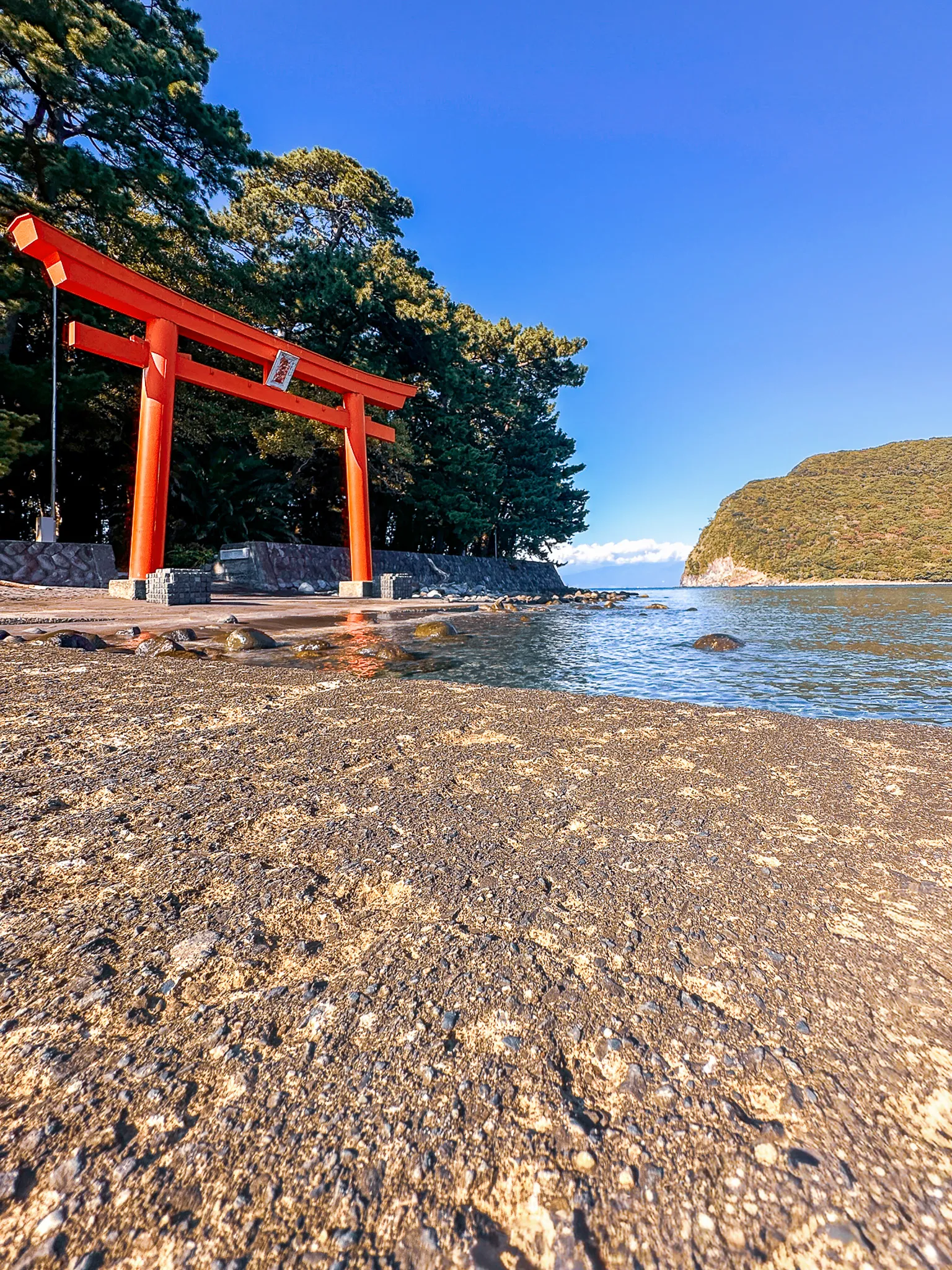 A vermilion torii standing on a shingle beach at the water's edge, pines behind it and a wooded headland across the clear bay under a deep blue sky