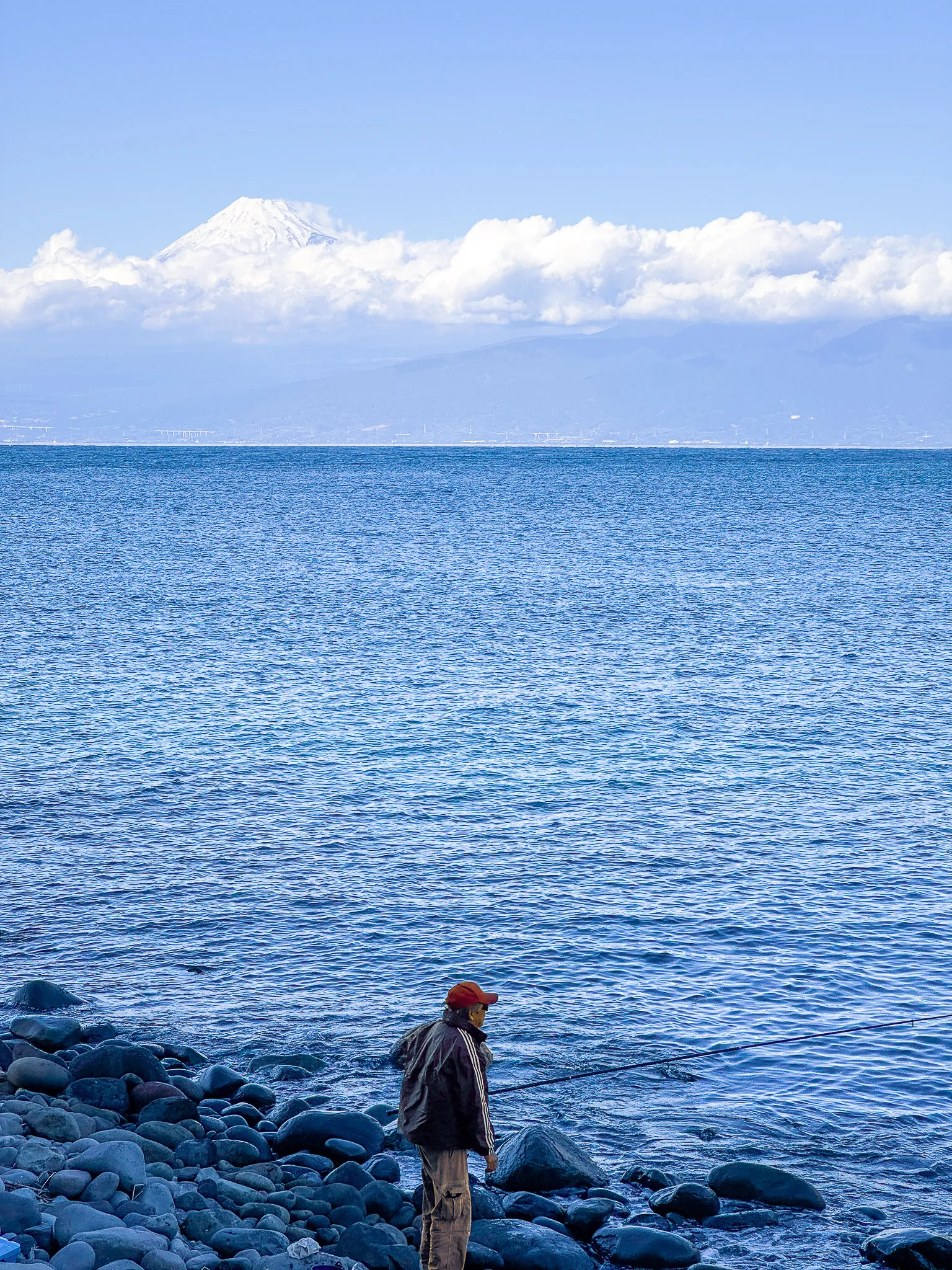 Mount Fuji snow-capped above a band of cloud across the water, and in the foreground a man in a red cap standing on a cobbled shore with a line out