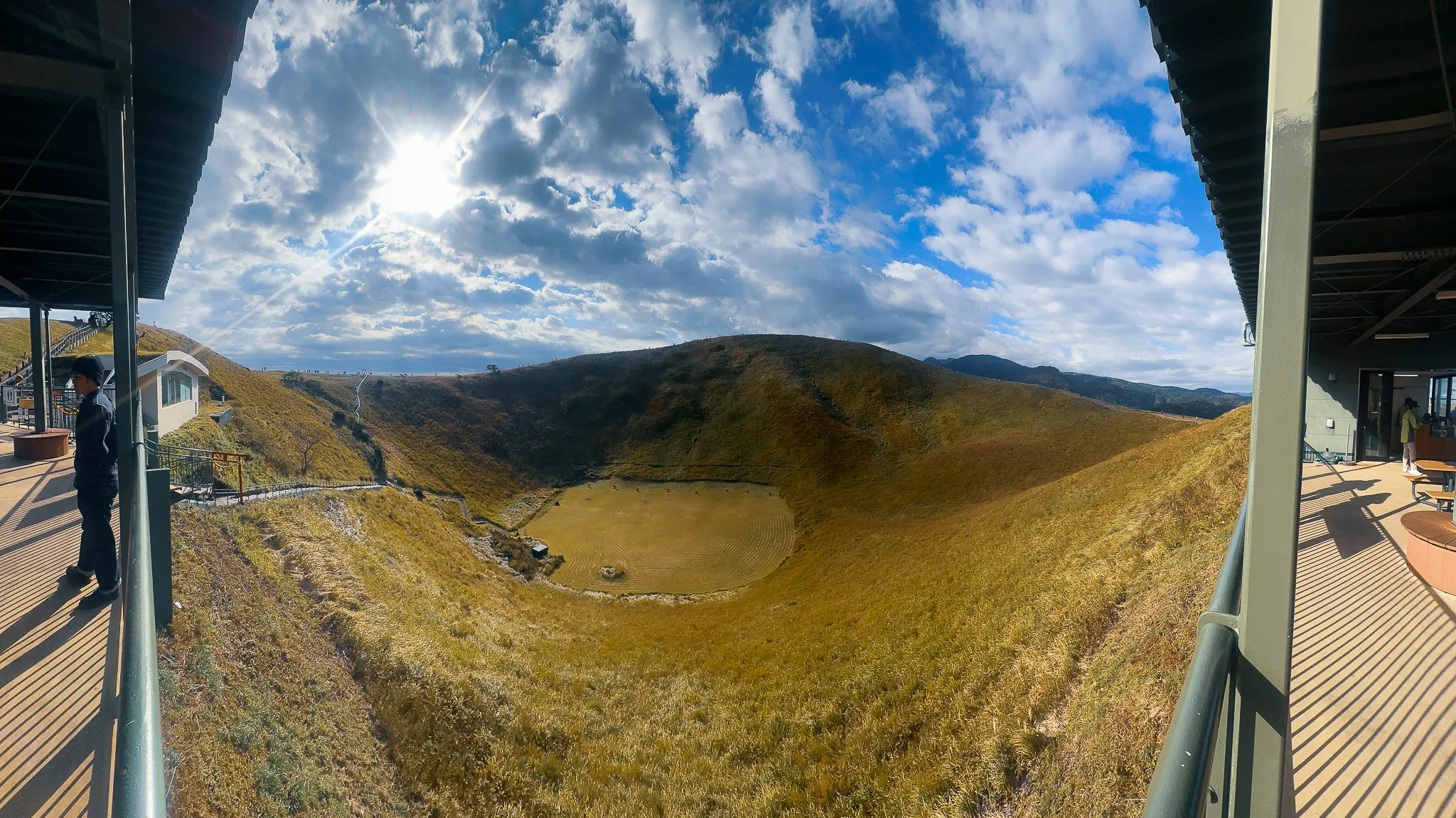 The grassed crater seen from the summit terrace, a flat green floor at the bottom of a steep bowl, a person standing at the rail on the left