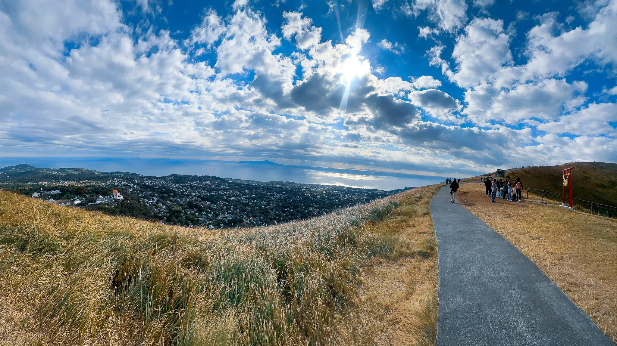 A wide panorama from the crater rim - a paved path curving away with walkers on it, dry grass falling to the town and the bay below, the sun breaking through a big cloud bank