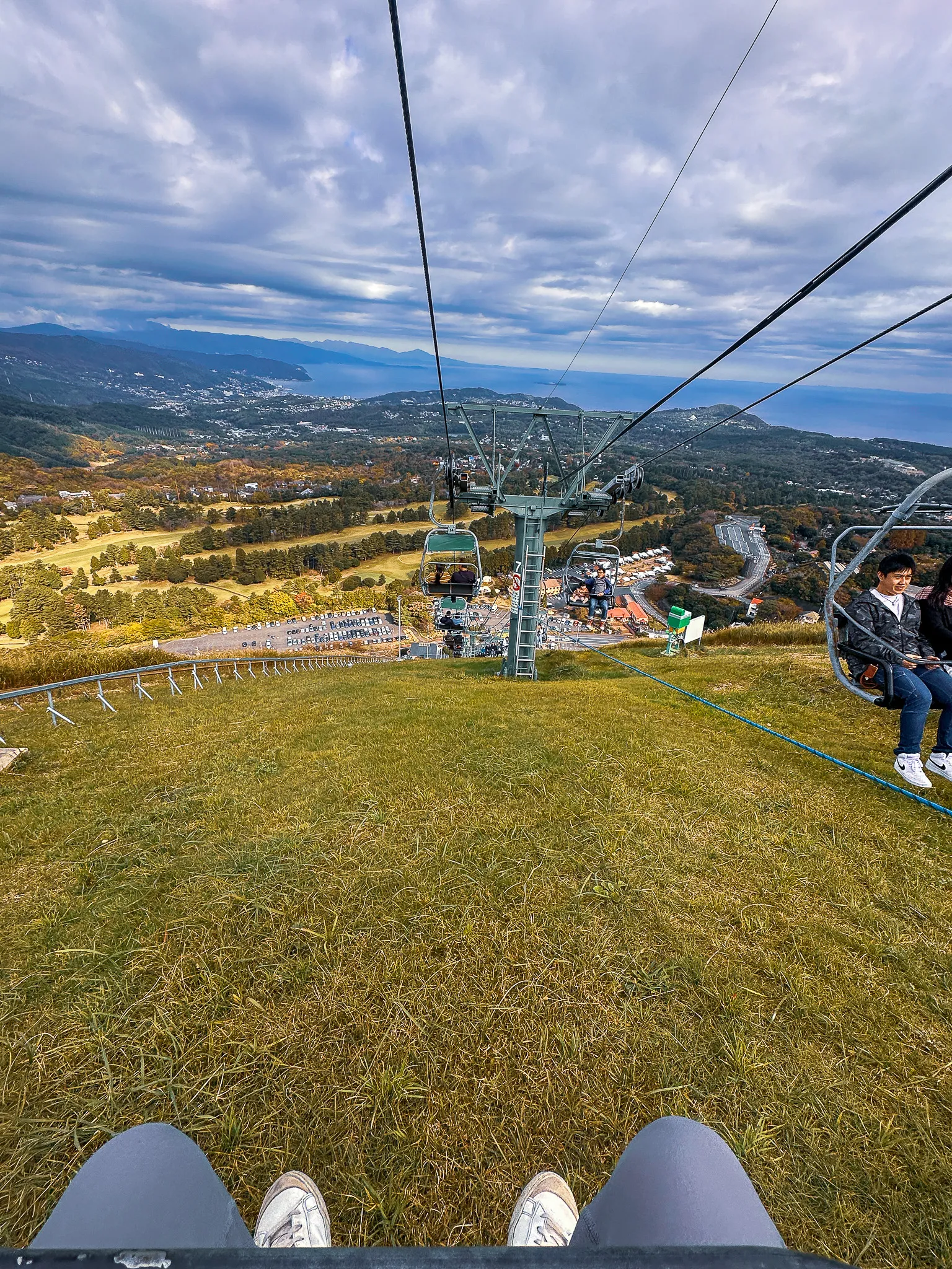 The view from a chairlift riding down a grass slope, my shoes in the frame, the town and the bay spread out below under heavy cloud
