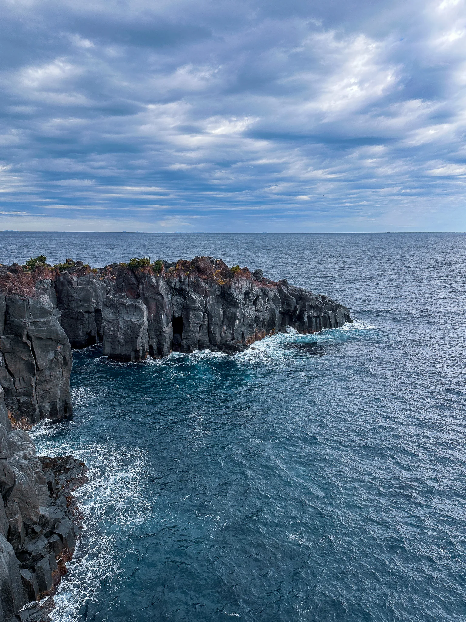Columns of dark lava cliff running out to a headland with a cave at its foot, surf breaking pale green over the shallows and a low grey cloud bank out to sea