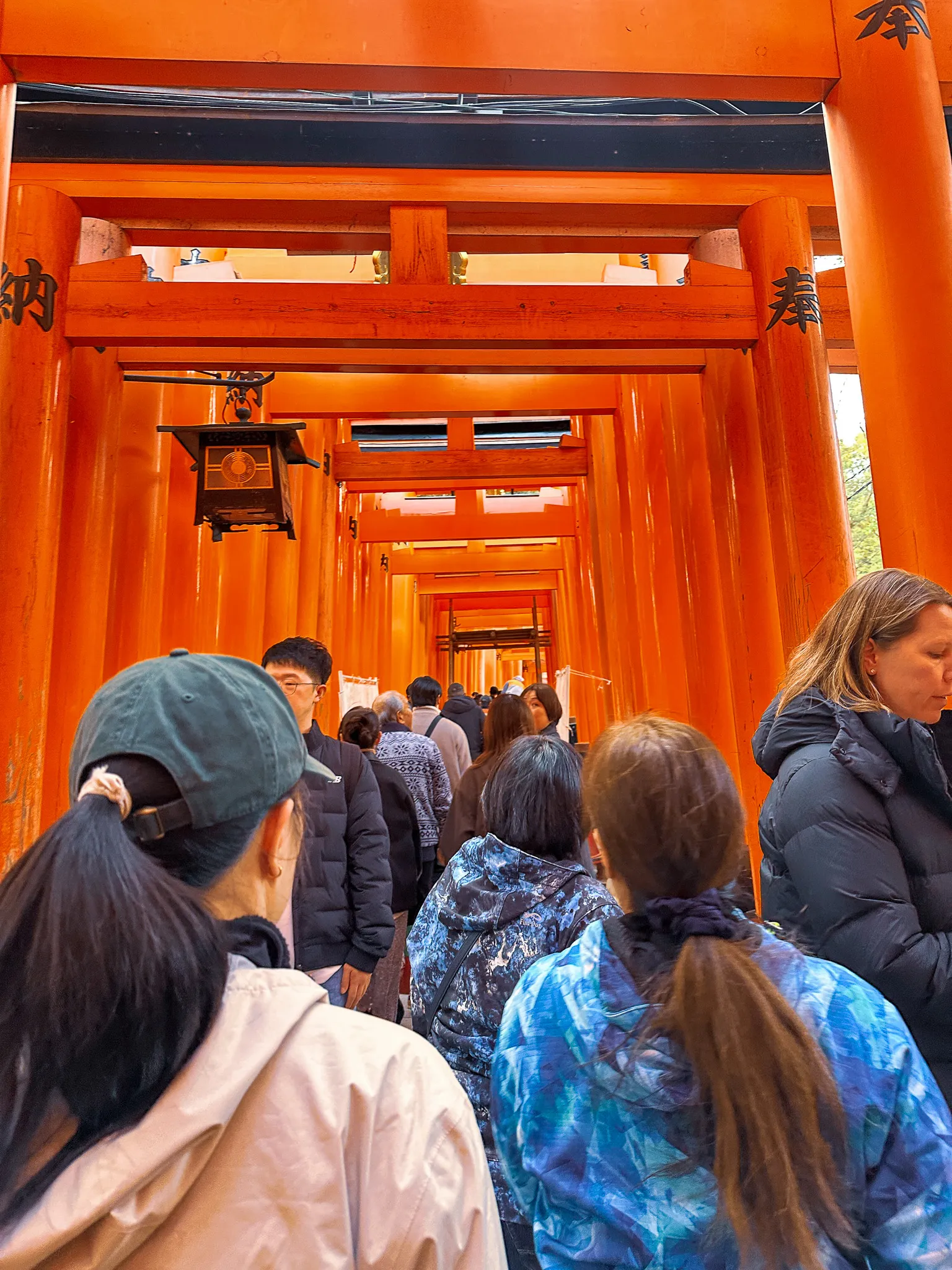 Inside the tunnel of torii at Fushimi Inari, packed with visitors walking in single file, a hanging bronze lantern and inscribed pillars overhead