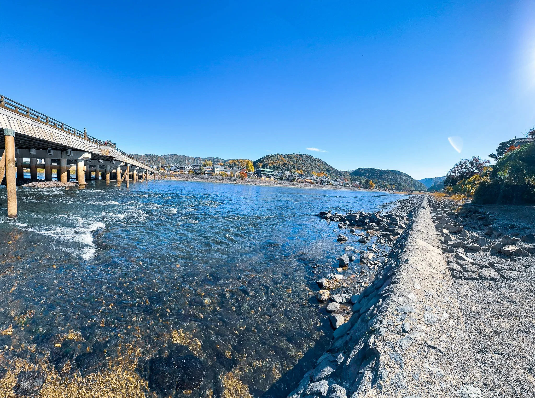 The Uji river running shallow and clear over stones, a long wooden bridge crossing out from the left and low wooded hills beyond