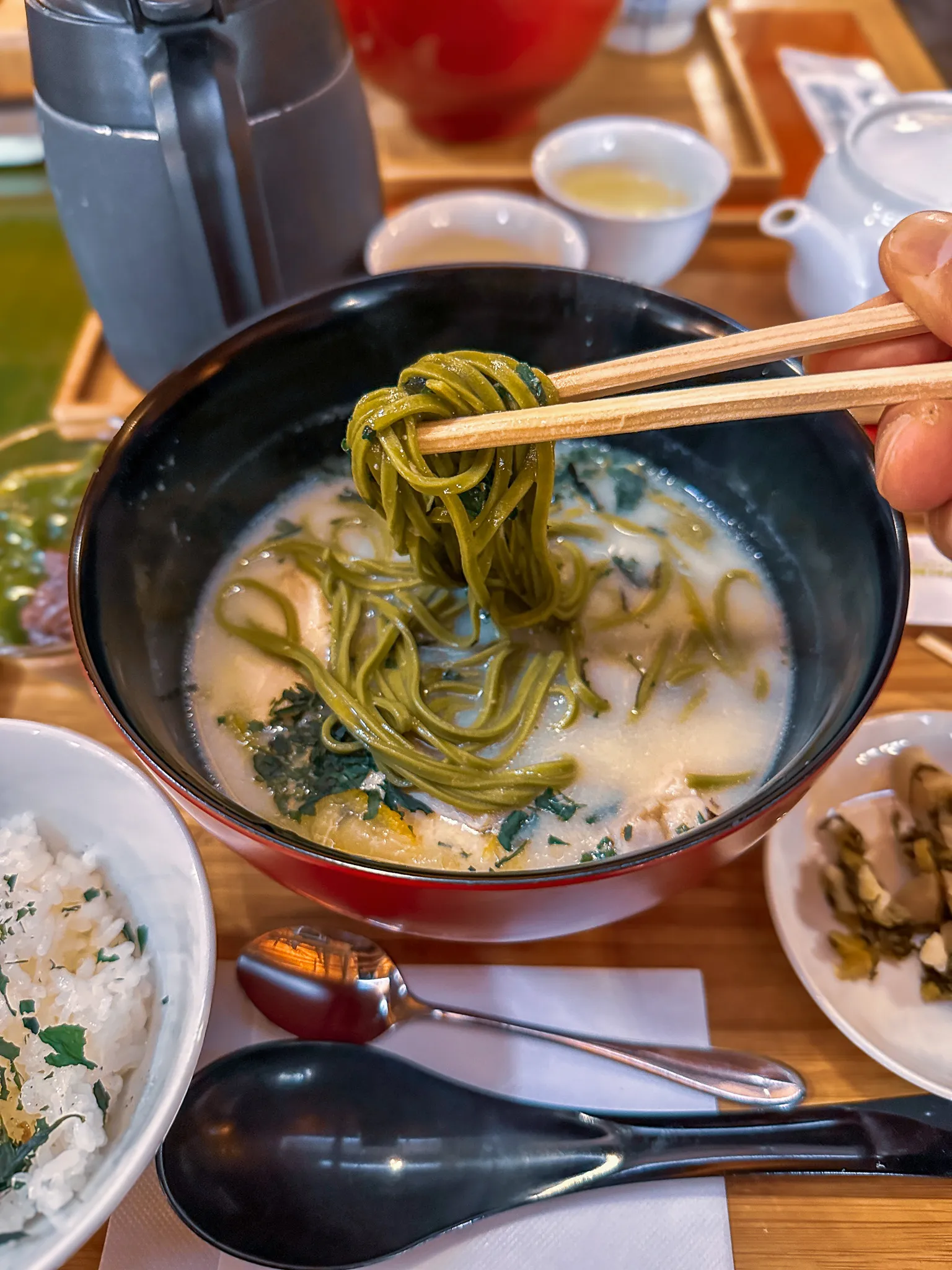 Green tea noodles lifted on chopsticks from a bowl of milky white broth in black and red lacquer, with rice, pickles and a pot of tea on the table around it