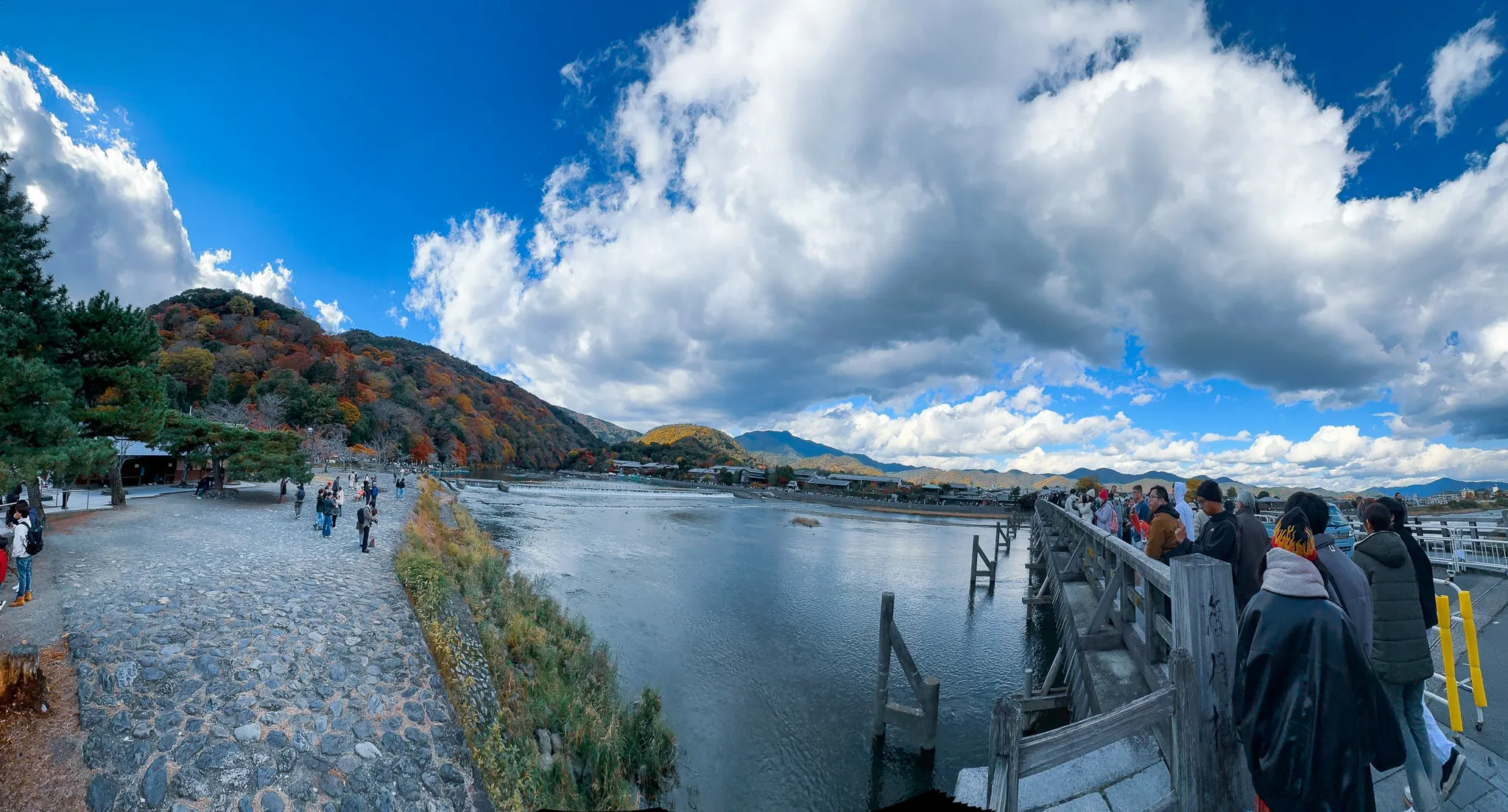 A wide panorama from the Togetsukyo bridge - the gravel path and hills on the left, the river running through the middle, and the bridge's wooden rail crowded with people on the right