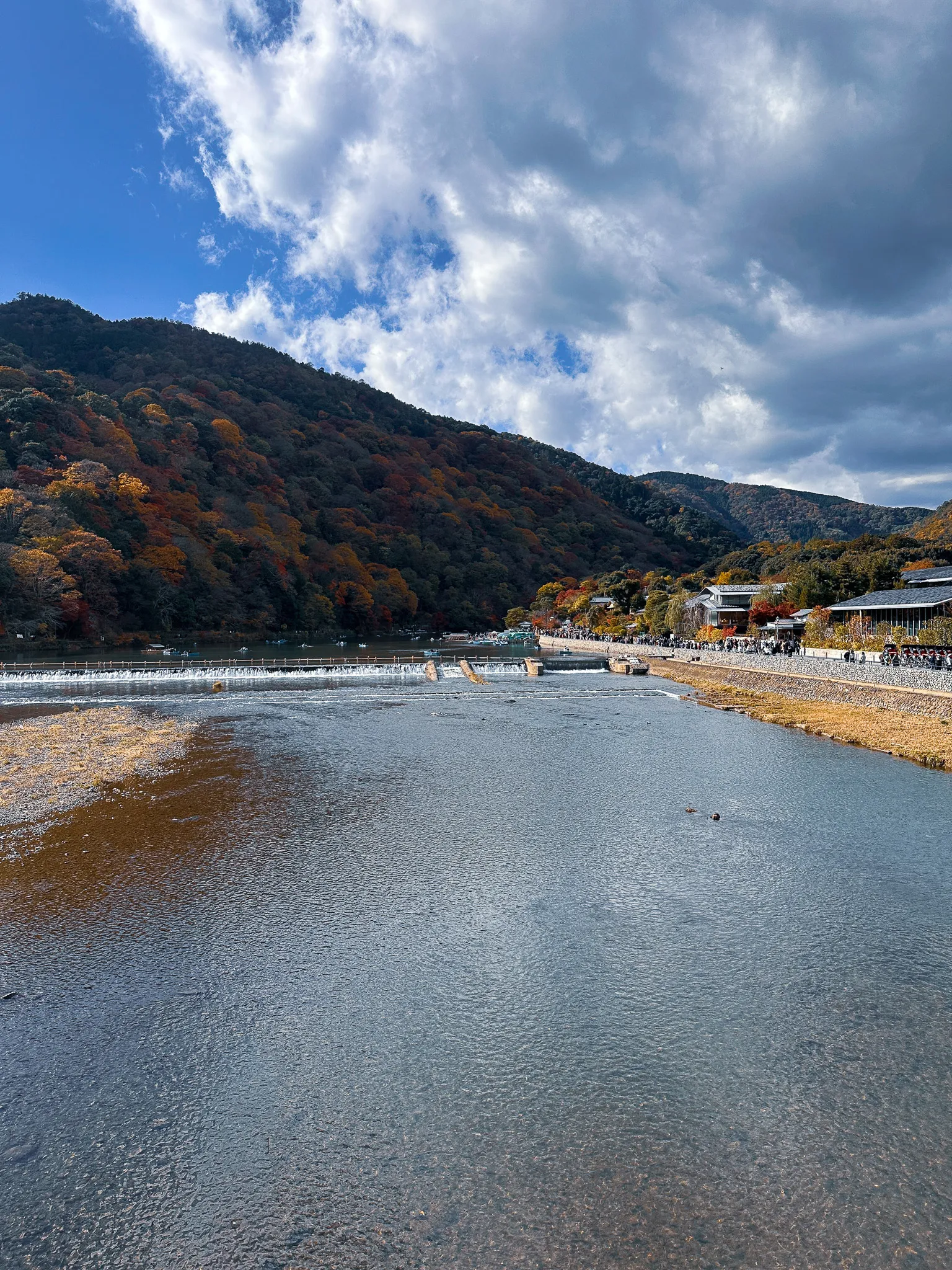 The river running wide and shallow away from the bridge, the wooded hillside on the left and the riverside path crowded with people on the right under big clouds